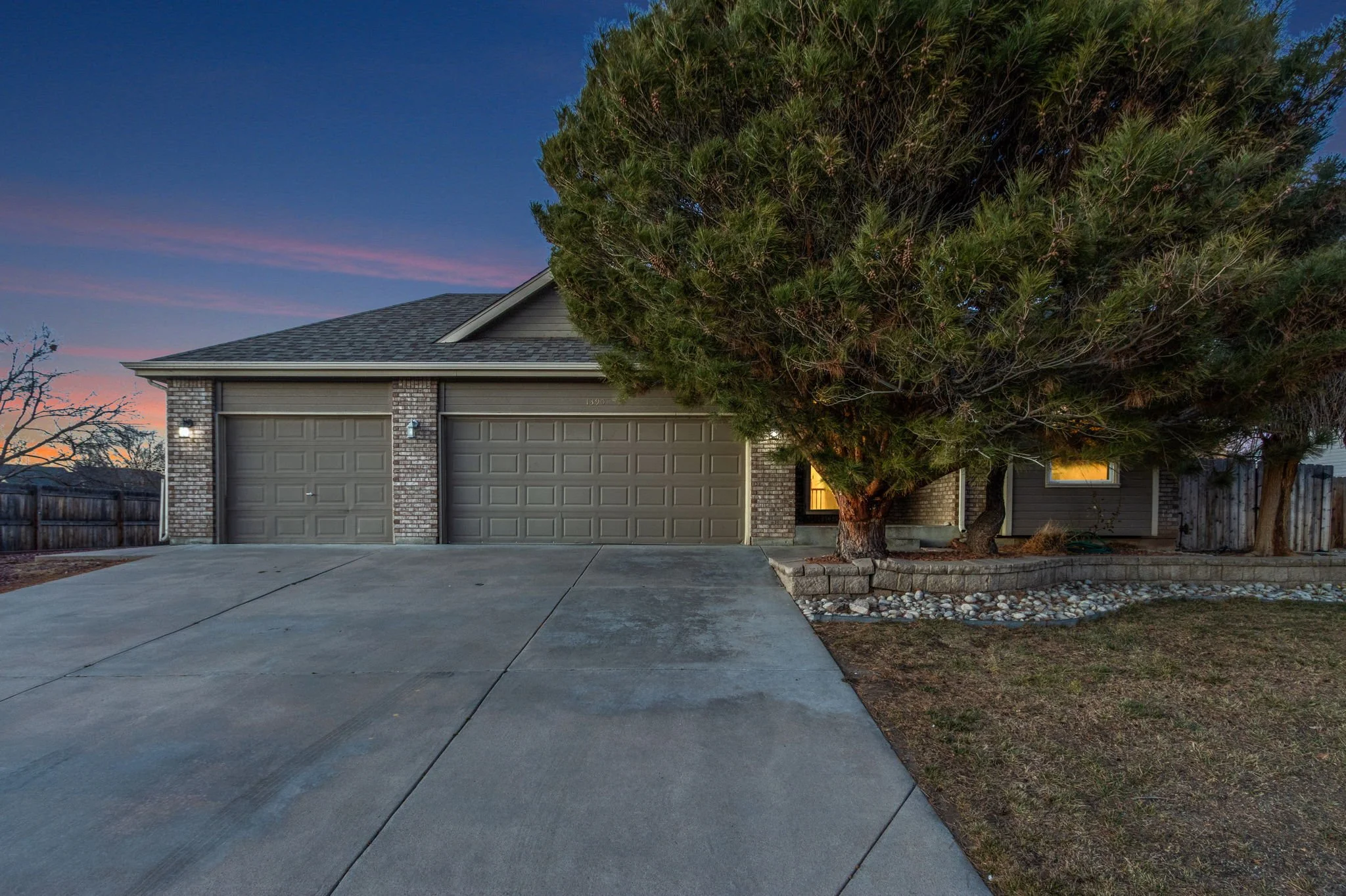 A residential house with a two-car garage, a large tree in front, and a evening sky with pink and blue hues.