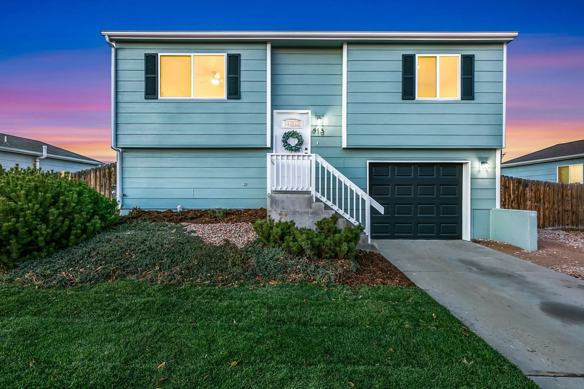 Front of a two-story blue house with a black garage door, white stairs and railing, two yellow-lit windows, and a wreath on the front door, with a colorful sunset sky in the background.
