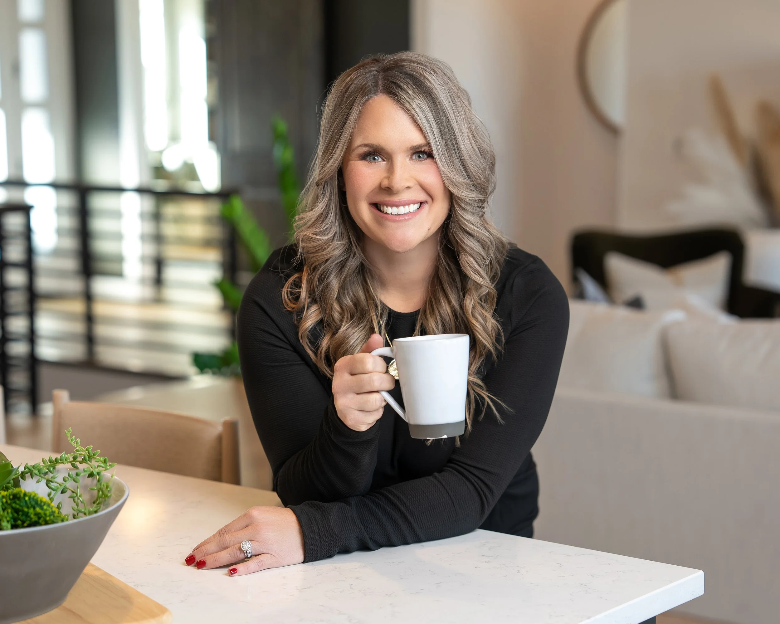 A woman with long gray hair and a bright smile, holding a white mug, sitting at a table in a well-lit room, with a plant visible in the foreground.