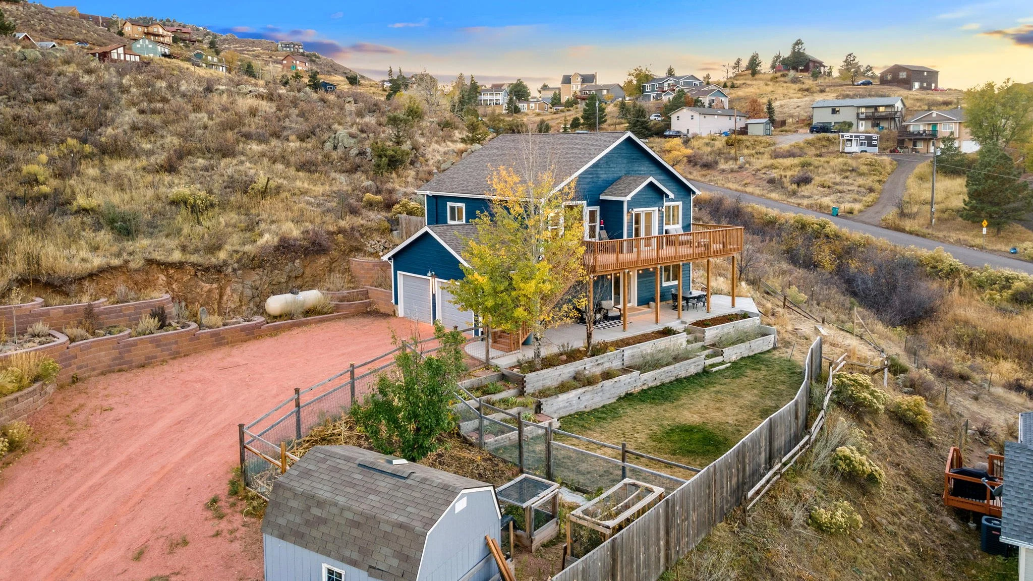 A blue two-story house with a deck, surrounded by a landscaped yard with a tree, fencing, and neighboring houses on a hillside with dry grass and shrubs under a partly cloudy sky.
