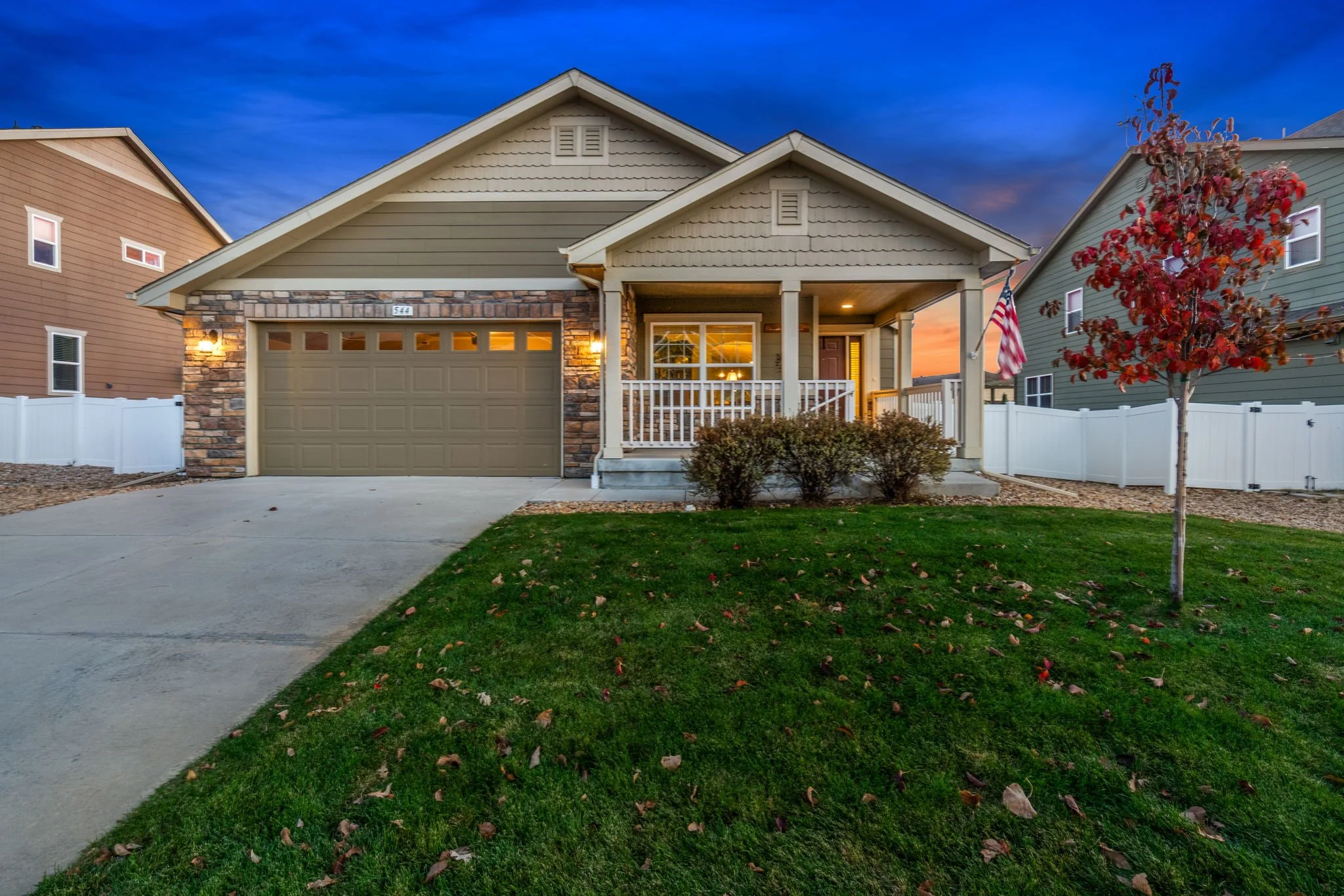 A house with a stone and siding exterior, a two-car garage, a front porch, and a small tree with red leaves in the yard during sunset.
