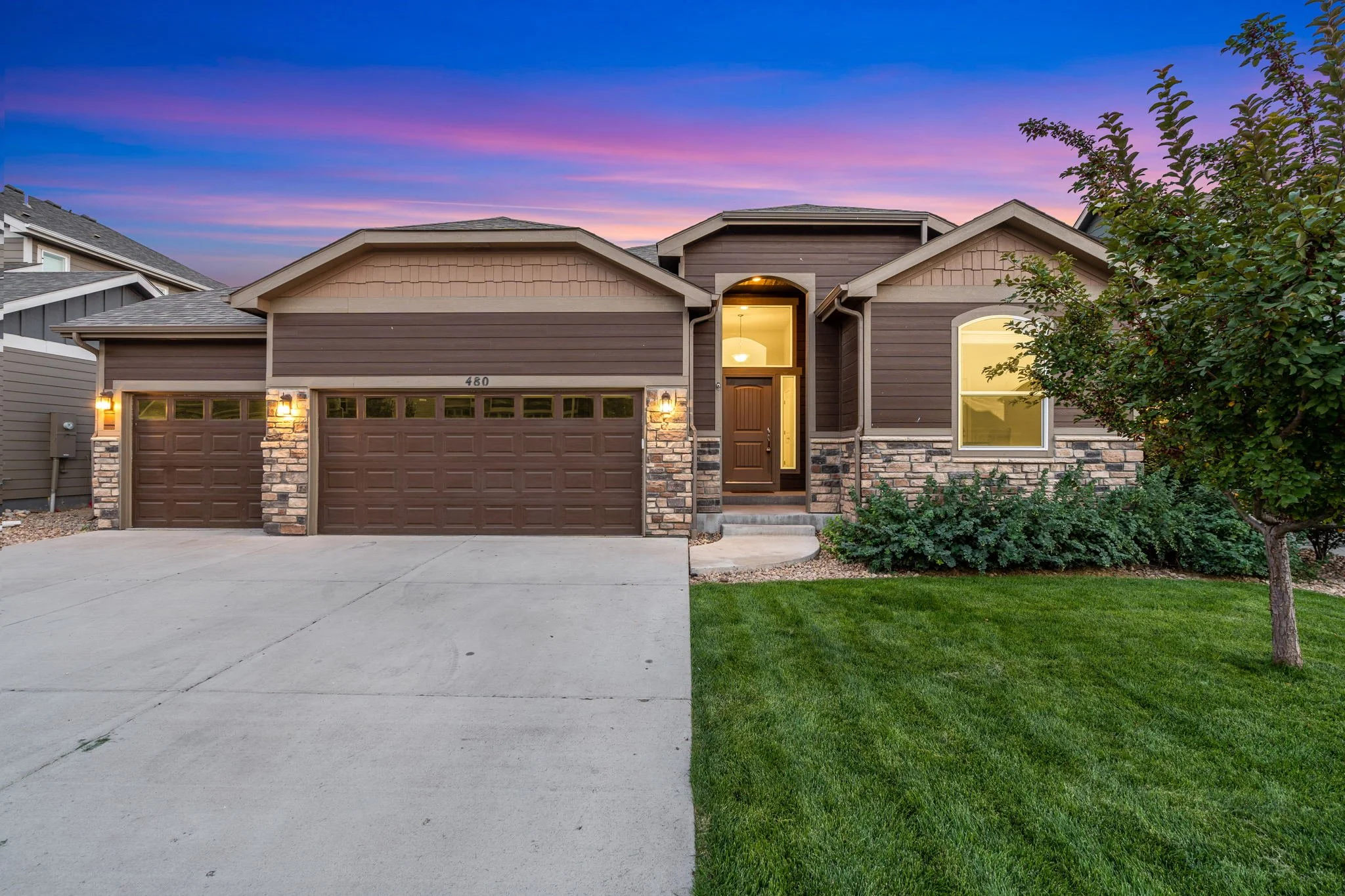 A modern two-story house with a two-car garage, stone accents, and a front door illuminated by exterior lights during sunset, with a green lawn and trees.