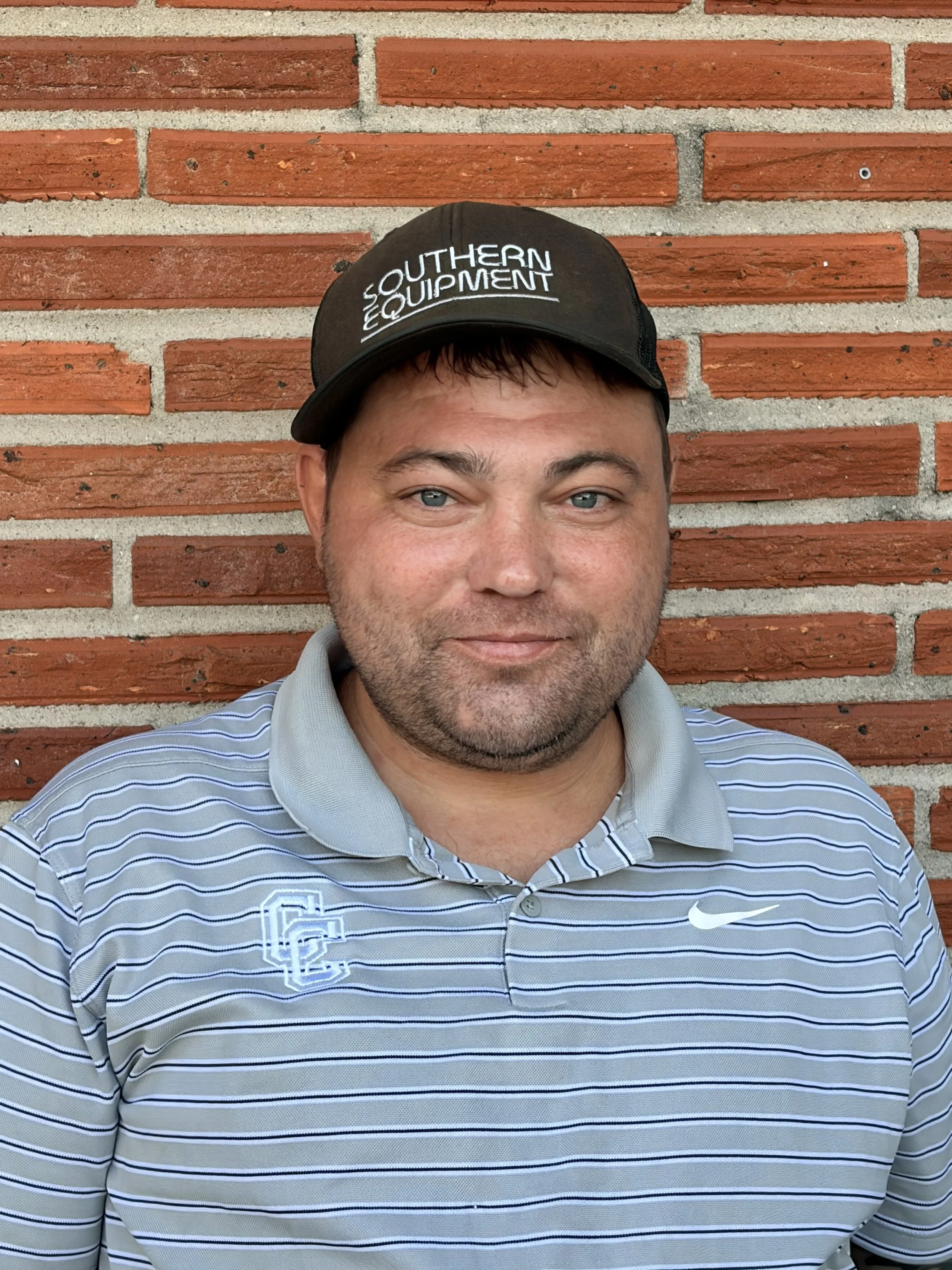 A man wearing a striped polo shirt and a black cap with white embroidered text 'Southern Equipment' stands in front of a red brick wall, looking at the camera with a slight smile.