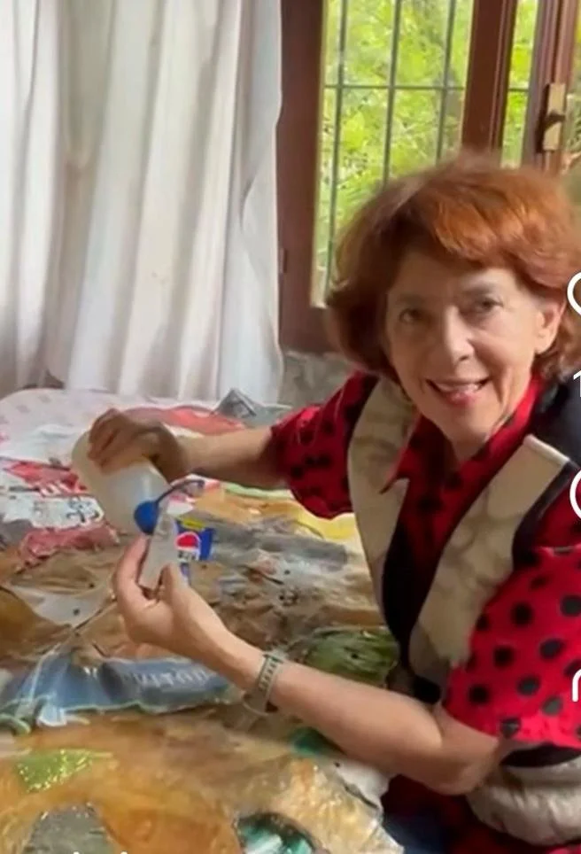 A woman with red hair smiling at the camera, sitting at a table with a blanket or cloth with various colorful prints, pouring liquid into a measuring cup, near a window with green foliage outside.