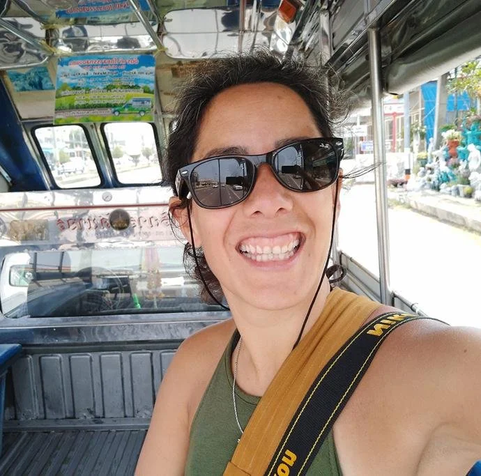 Woman with dark sunglasses smiling in a tuk-tuk, wearing a green top and a yellow strap bag, on a busy street.