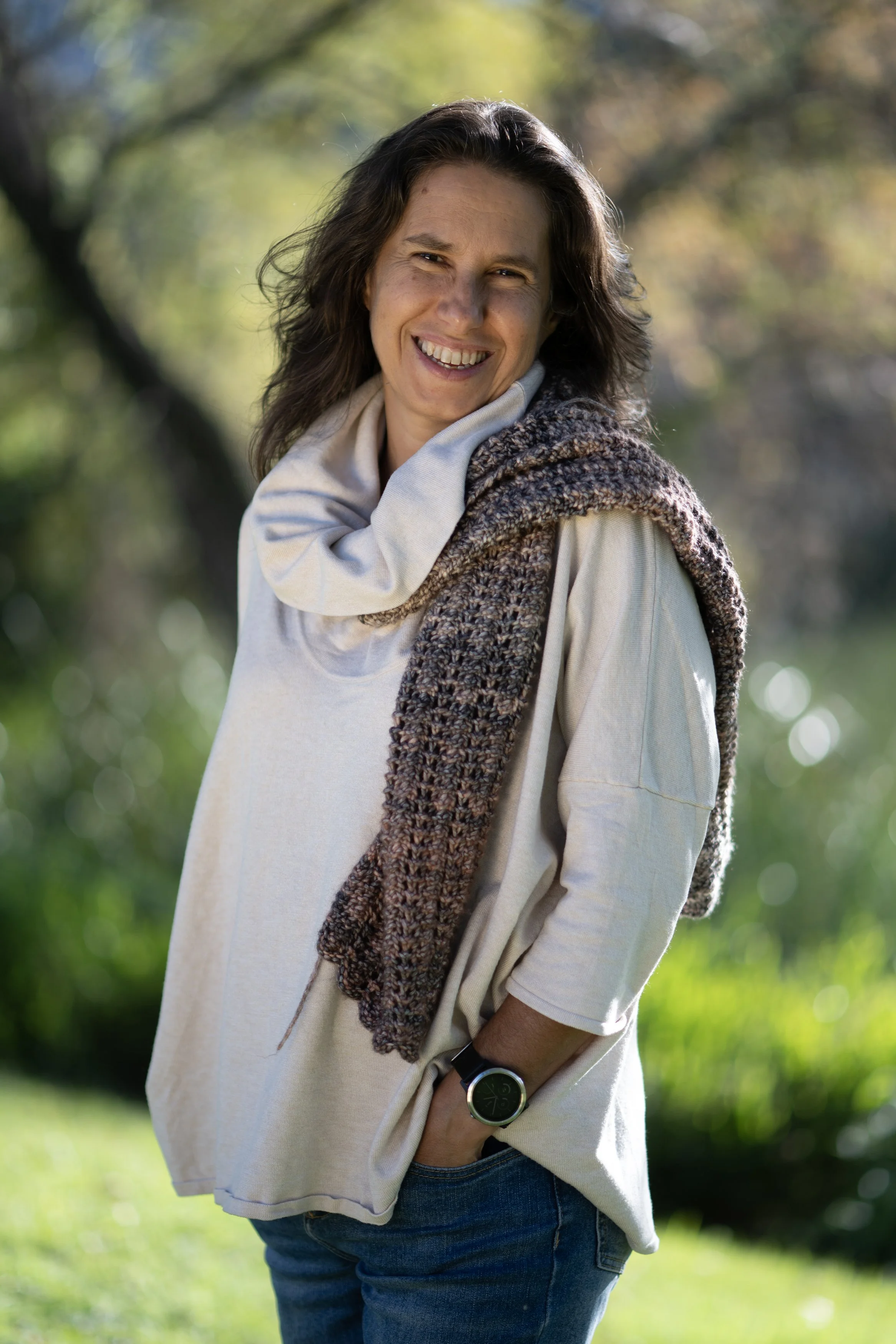 A woman with dark, wavy hair smiling outdoors in a park, wearing a beige oversized sweater, a knitted scarf, and a wristwatch.