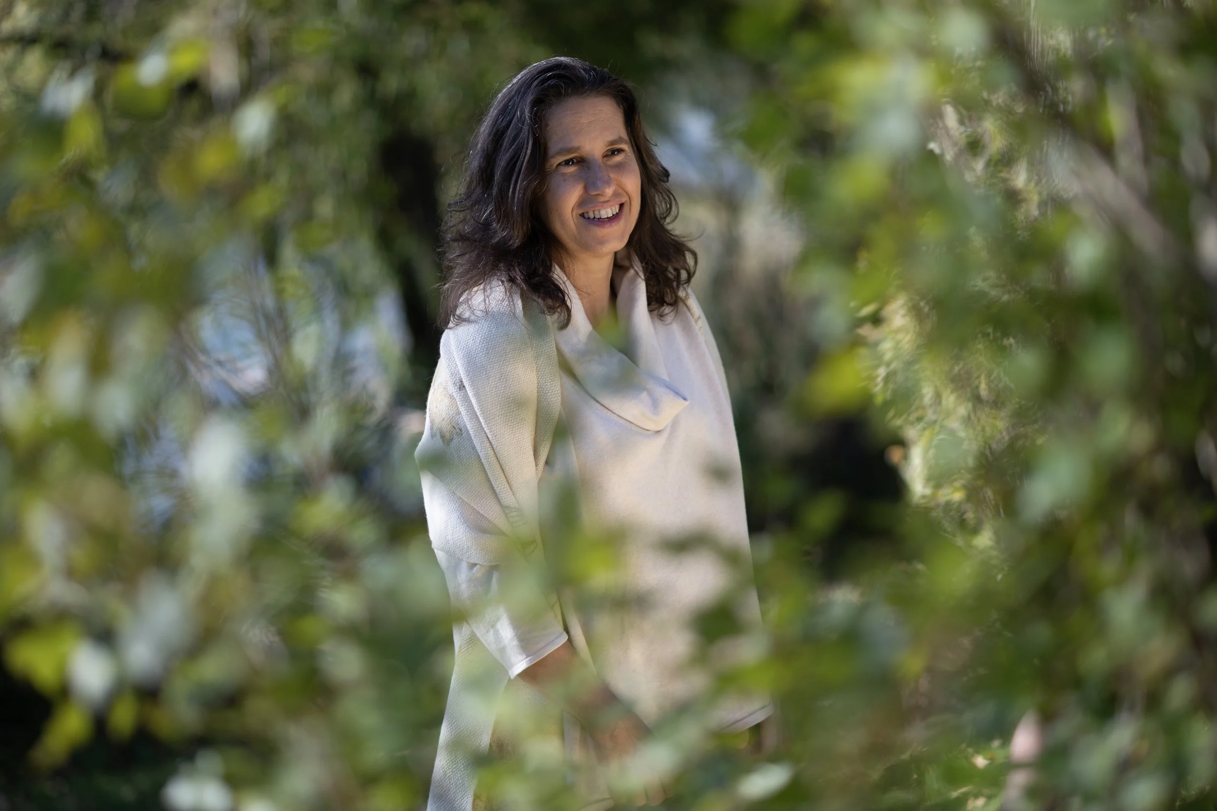 A woman with dark wavy hair smiling outdoors, surrounded by green foliage.