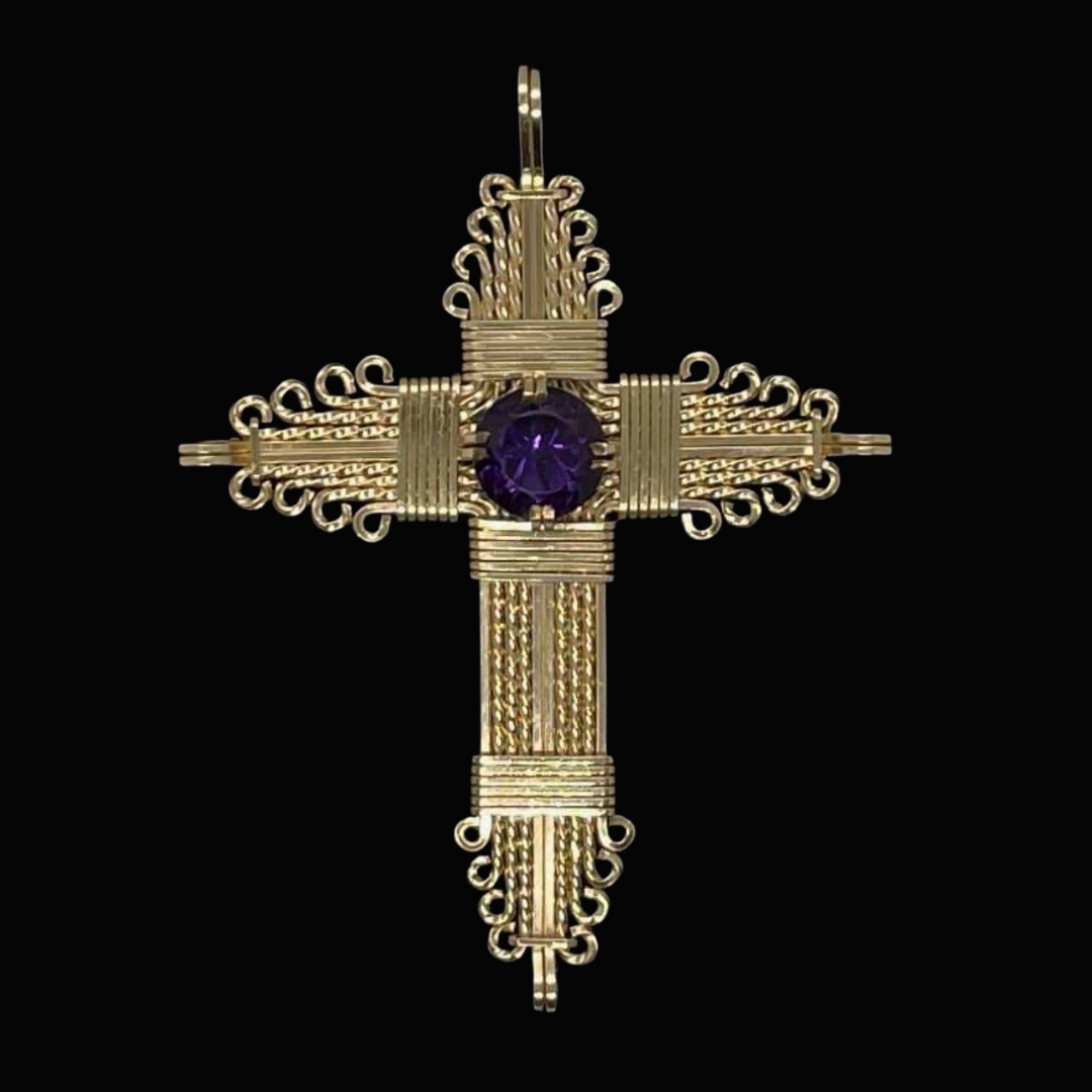 Gold cross pendant with intricate scrollwork and a purple gemstone at the center, set against a black background.