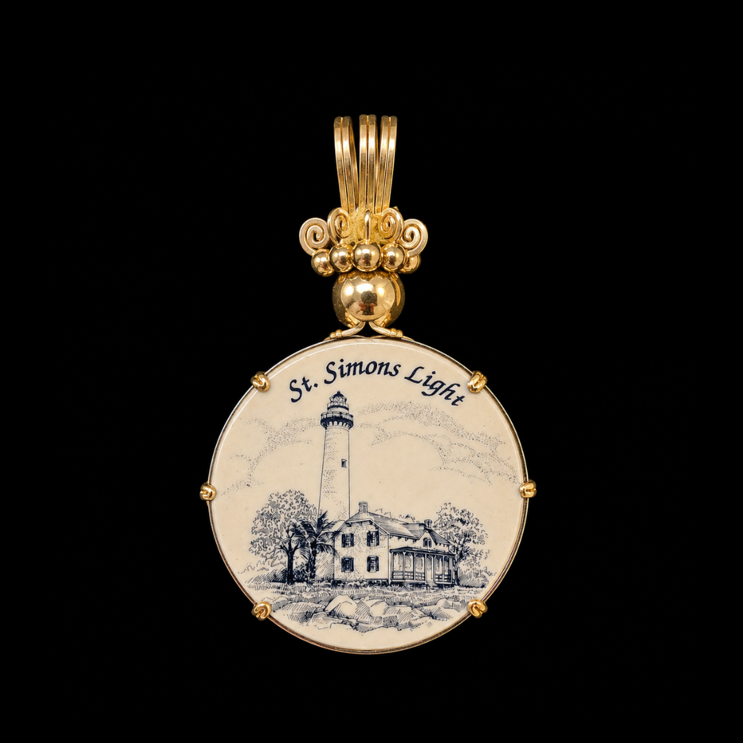 A gold-colored pendant with a circular white background featuring an illustration of a lighthouse, a house, and trees. The pendant has the words "St. Simons Island" written on it.