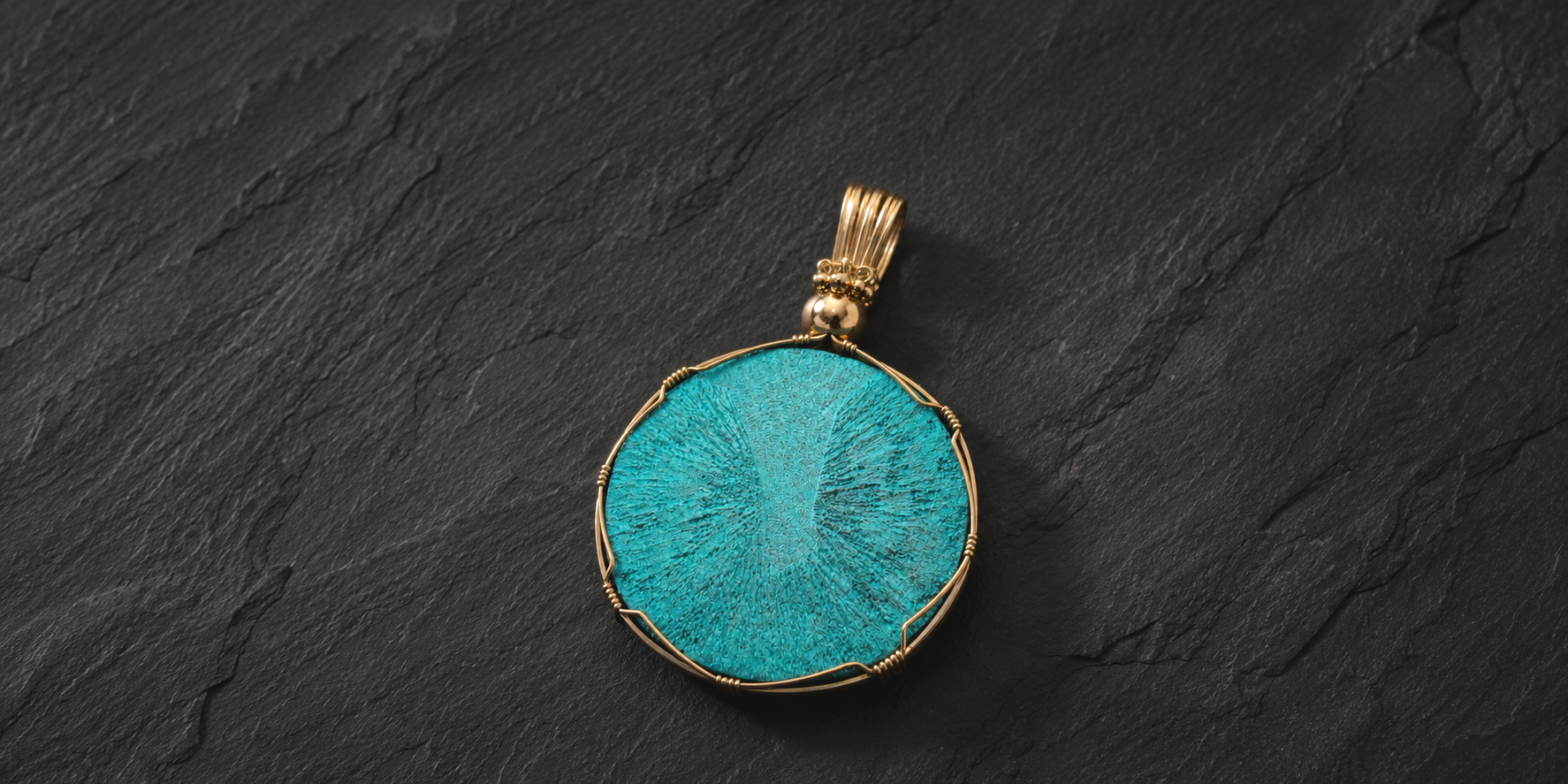 Gold pendant with a round, turquoise stone on a black textured surface.