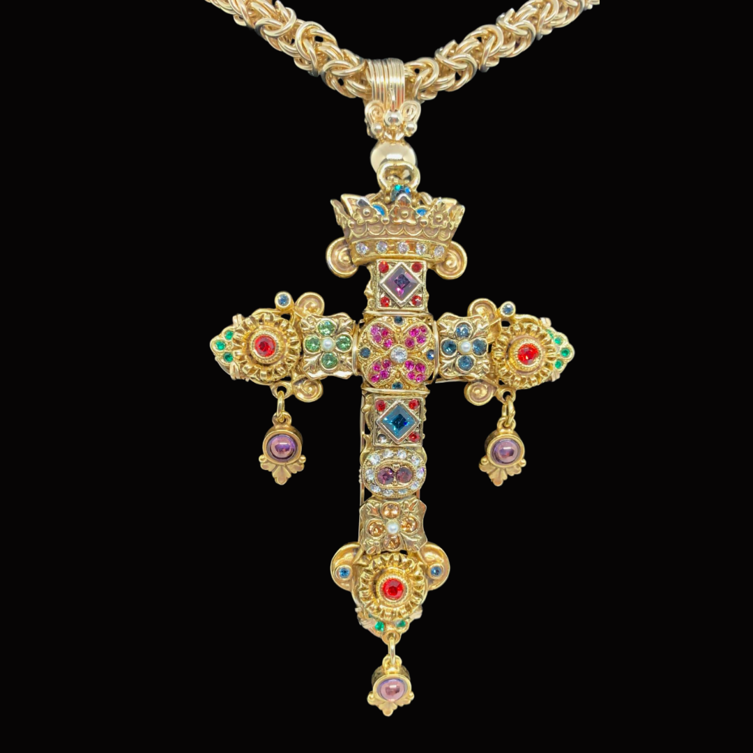 Gold chain necklace with a large ornate gold cross pendant adorned with colorful gemstones and small dangling ornaments, hanging against a black background.