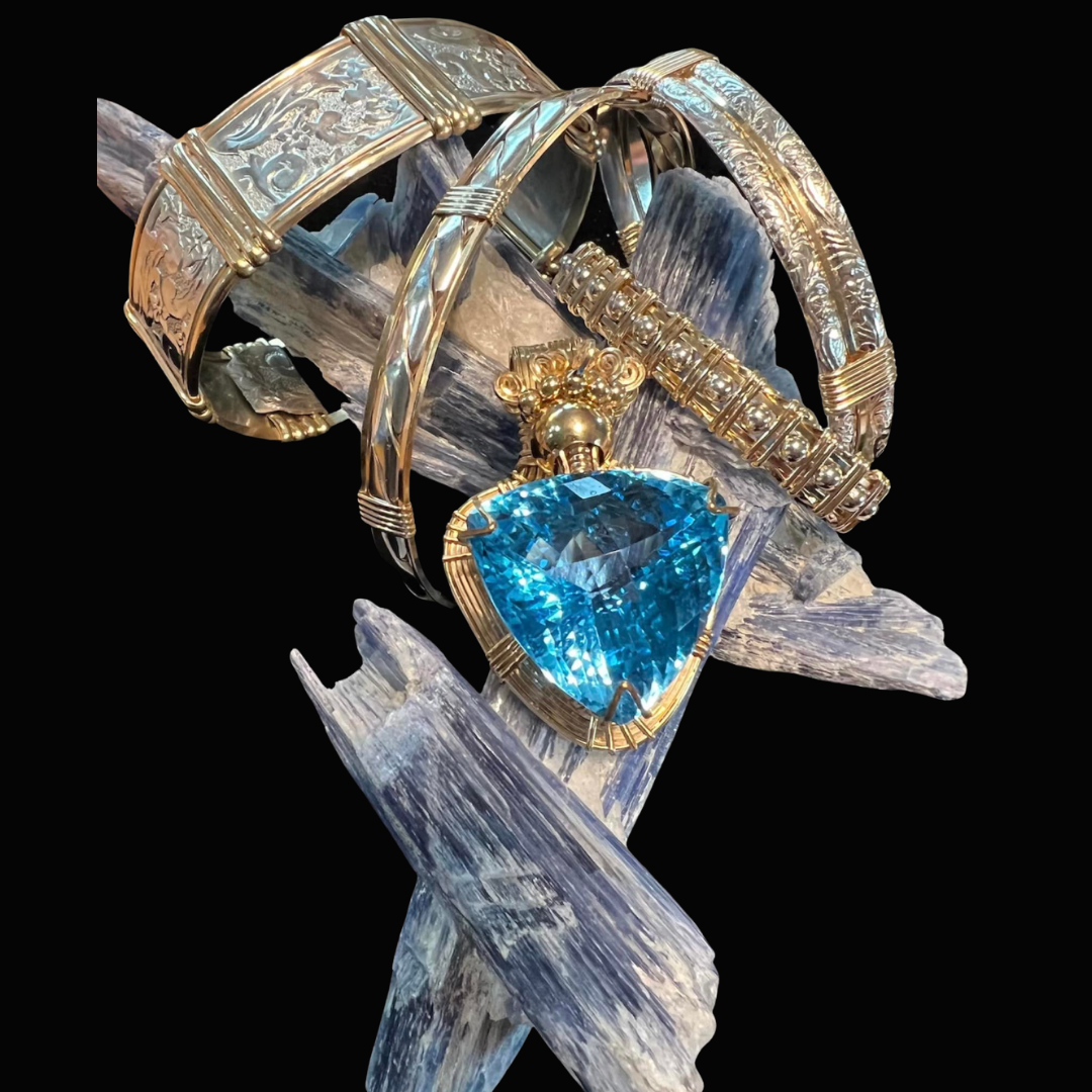 Jewelry including a large triangular blue gemstone ring and several gold and silver bracelets with detailed engravings, displayed on a piece of selenite crystal.