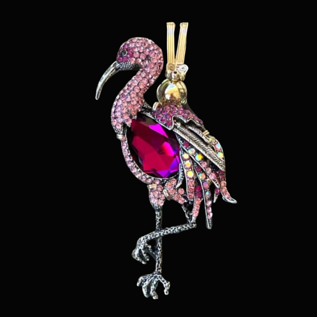A decorative brooch in the shape of a flamingo, adorned with pink and purple crystals and a large red gemstone on its body.