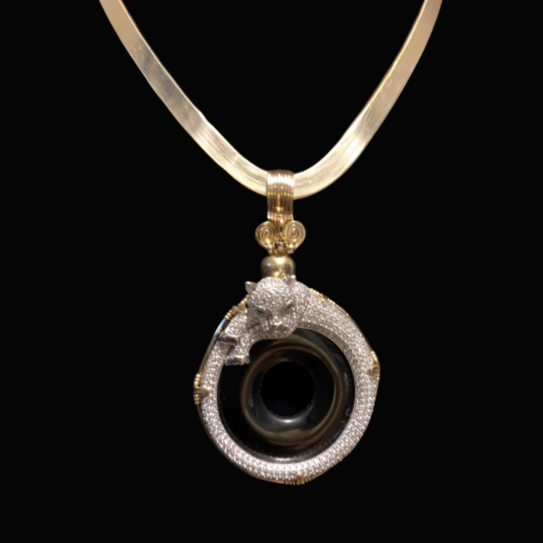 A close-up of a pendant on a necklace featuring a lion's head made of diamonds and a black circular gemstone or camera lens, with a gold setting and chain, against a black background.