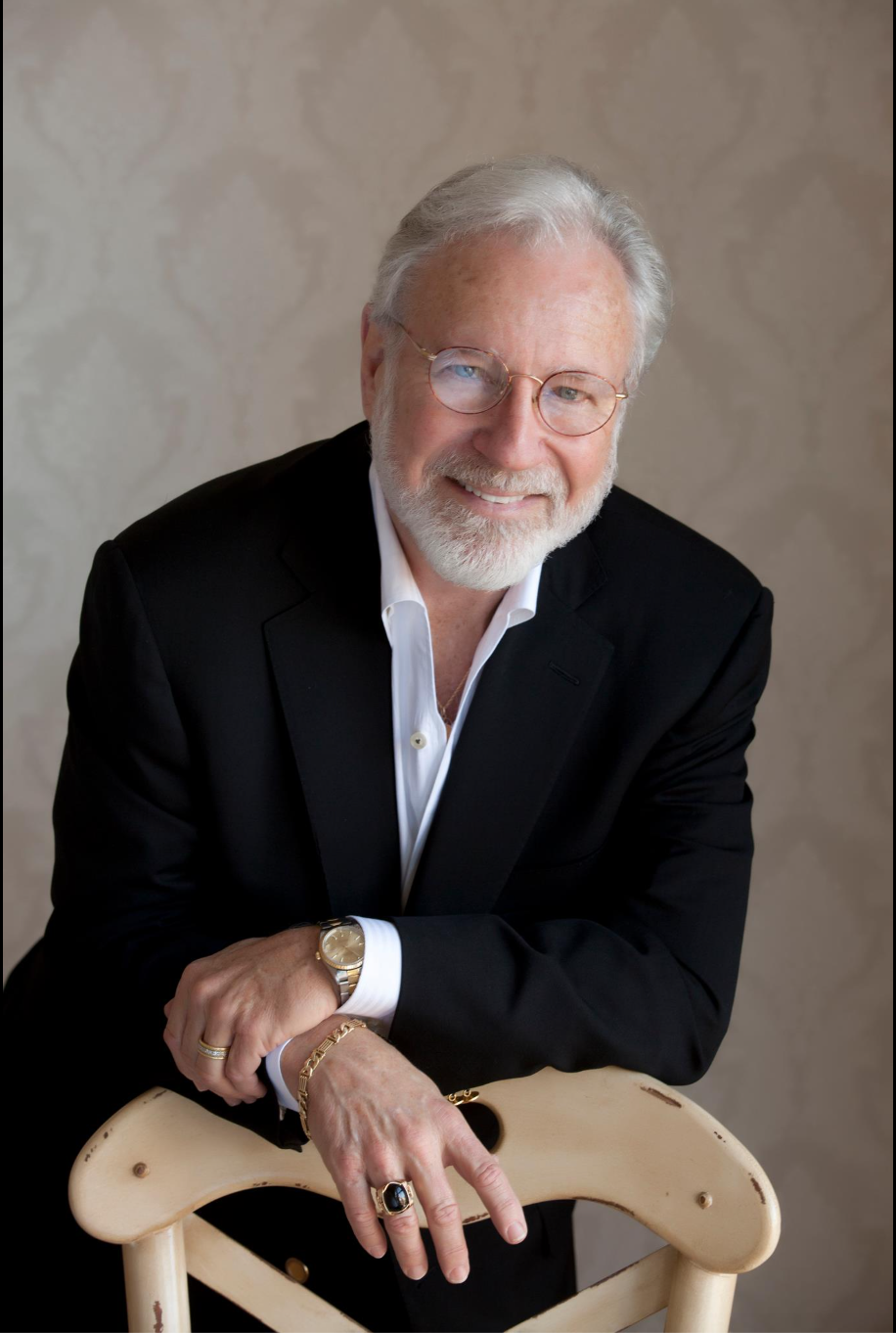 An older man with white hair and facial hair, wearing glasses, a black suit, and a white shirt, smiling at the camera while leaning on a wooden chair.