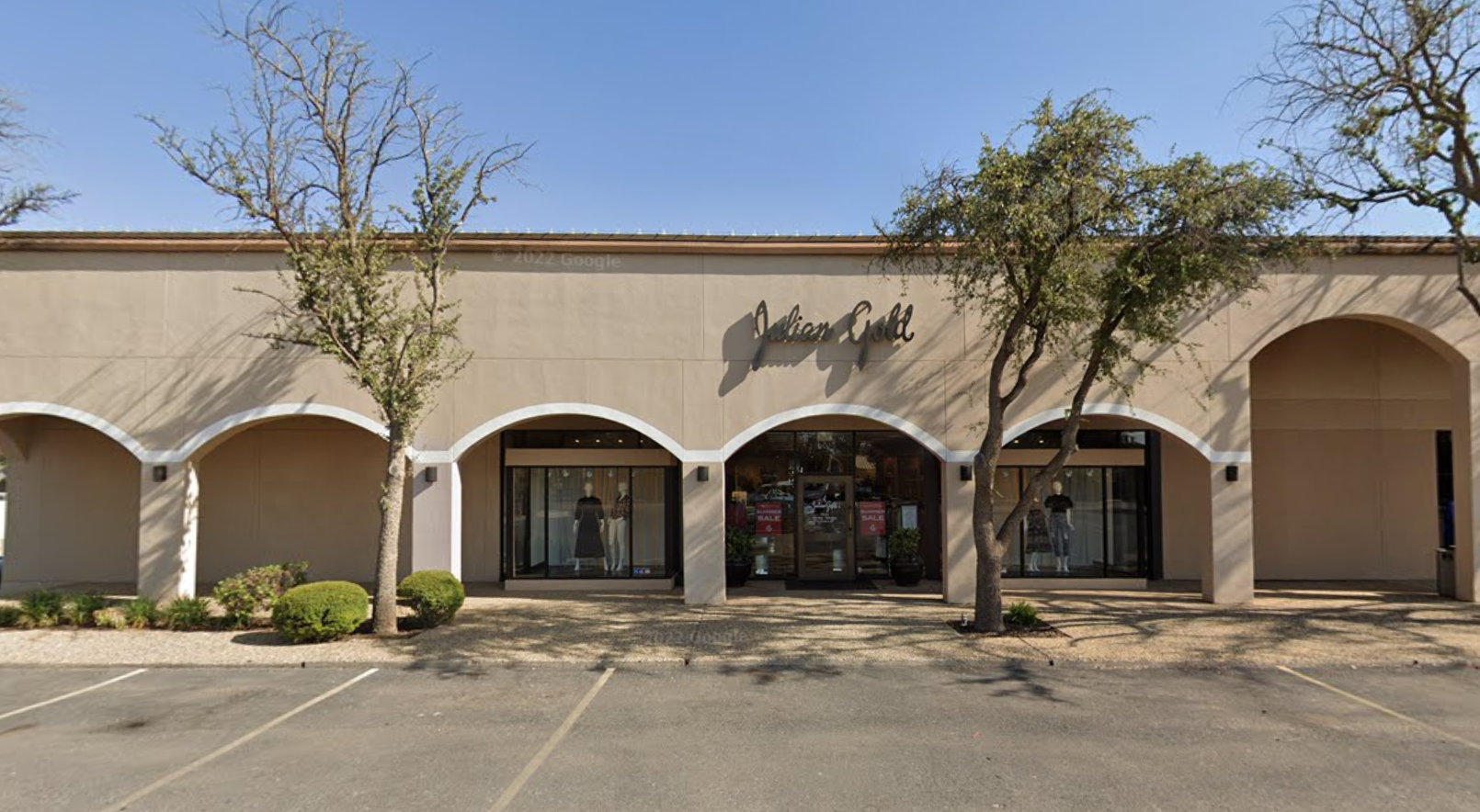 Exterior view of a jewelry store named Julian Gold with large glass windows, a beige facade, three trees in front, and a parking lot.