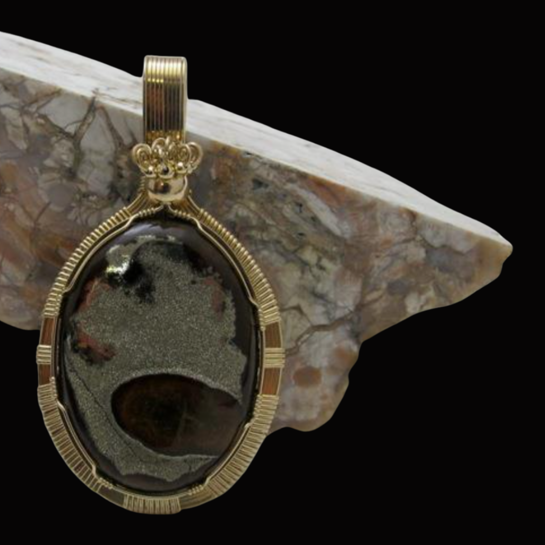 Gold pendant with black and gray patterned oval stone, hanging against a black background with a marble surface.