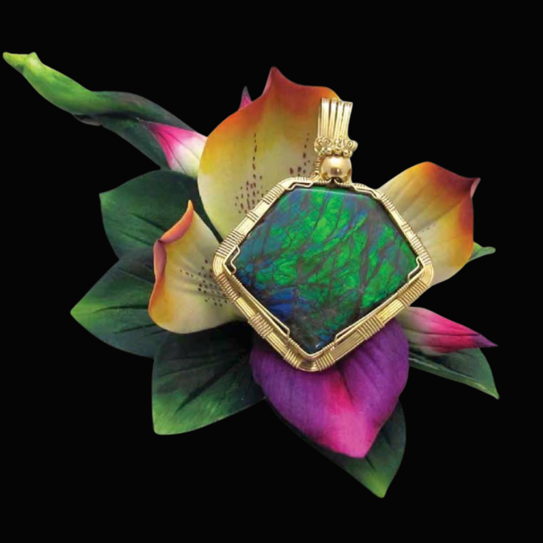 A large, iridescent gemstone set in a gold frame and pendant, resting on a colorful flower with pink, purple, yellow, and green petals and leaves against a black background.