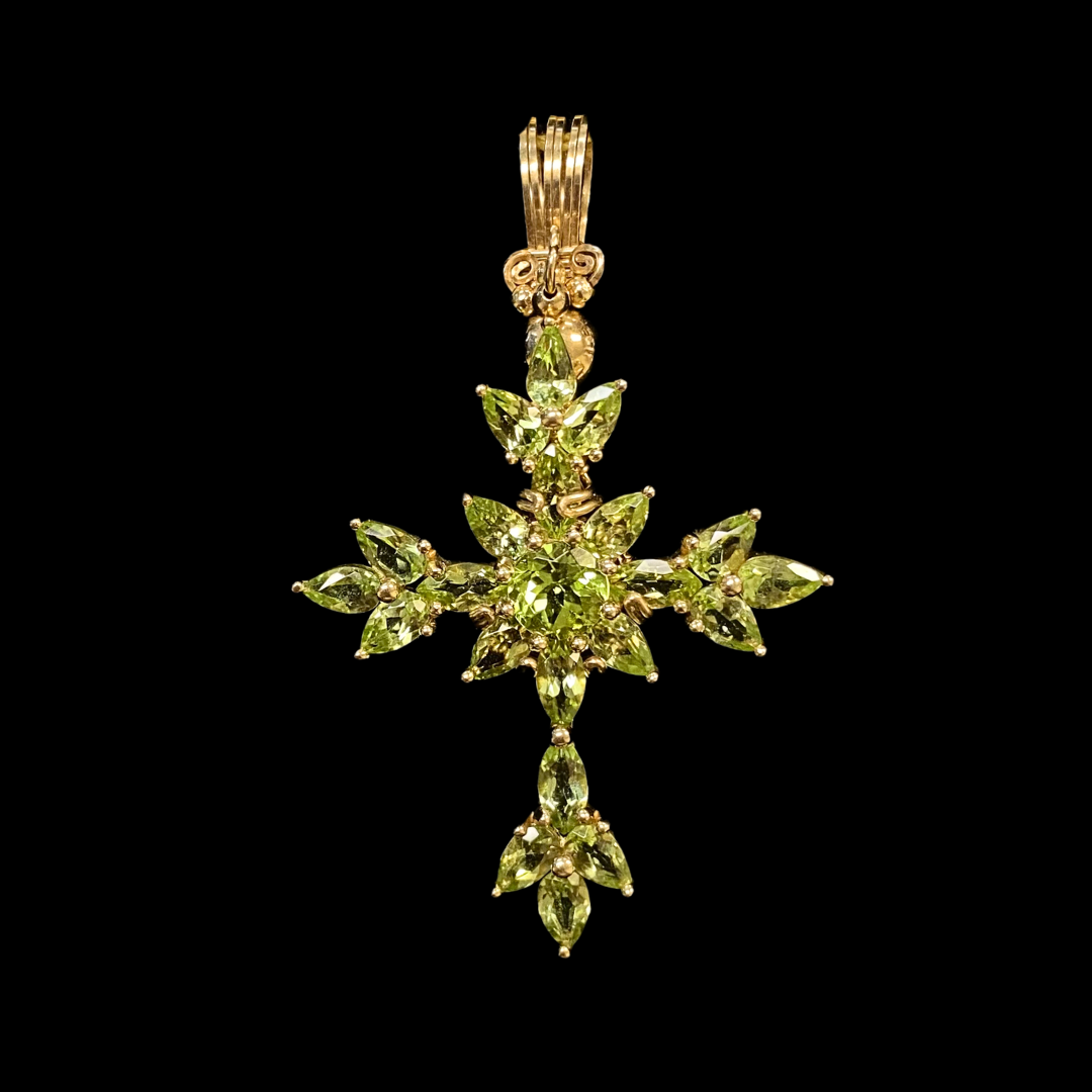 Gold cross pendant adorned with green gemstones and a gold top bail, set against a black background.