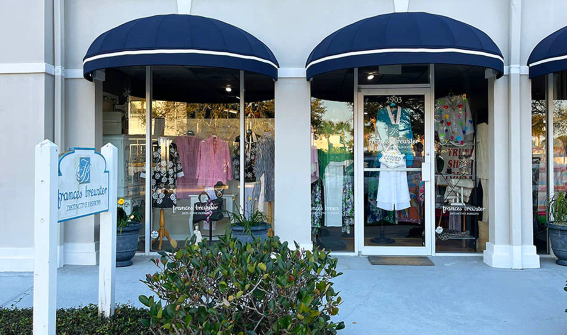 Front of a boutique clothing store with a glass window display showcasing various clothing items and a mannequin. The store has a white exterior with blue awnings and a white sign with the store's name, in blue lettering.