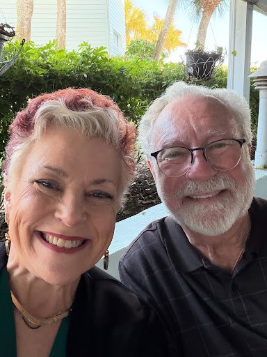 A smiling elderly woman with short gray and pink hair taking a selfie with an elderly man with white hair and glasses outdoors on a porch, with greenery and a house in the background.
