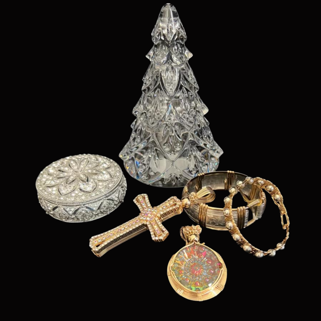Set of decorative jewelry and ornaments including a glass Christmas tree, a round silver jewelry box with floral detailing, a gold cross pendant with pink gemstones, two gold and pearl bracelets, and a circular pendant with a colorful design, all dis