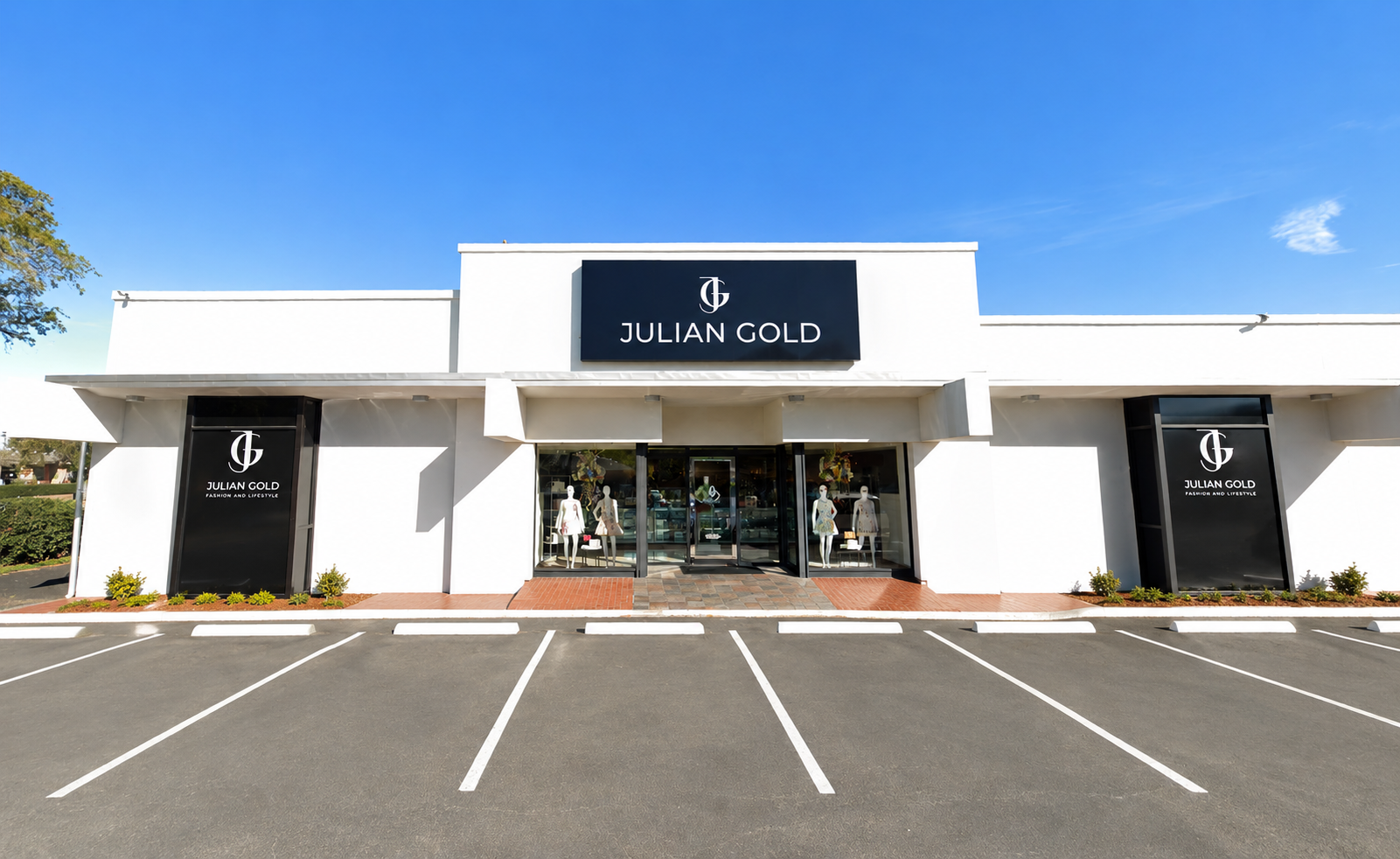 Front view of a white retail store called Julian Gold with large signs and mannequins displaying clothing in the windows, parking lot in front, under a clear blue sky.