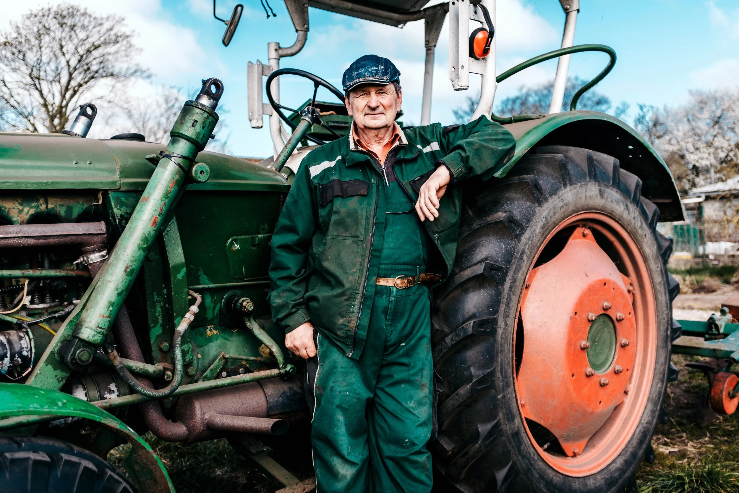 Älterer Mann in grüner Arbeitskleidung mit Baseballkappe lehnt an einem alten, grünen Traktor im Freien bei blauem Himmel und Bäumen im Hintergrund.