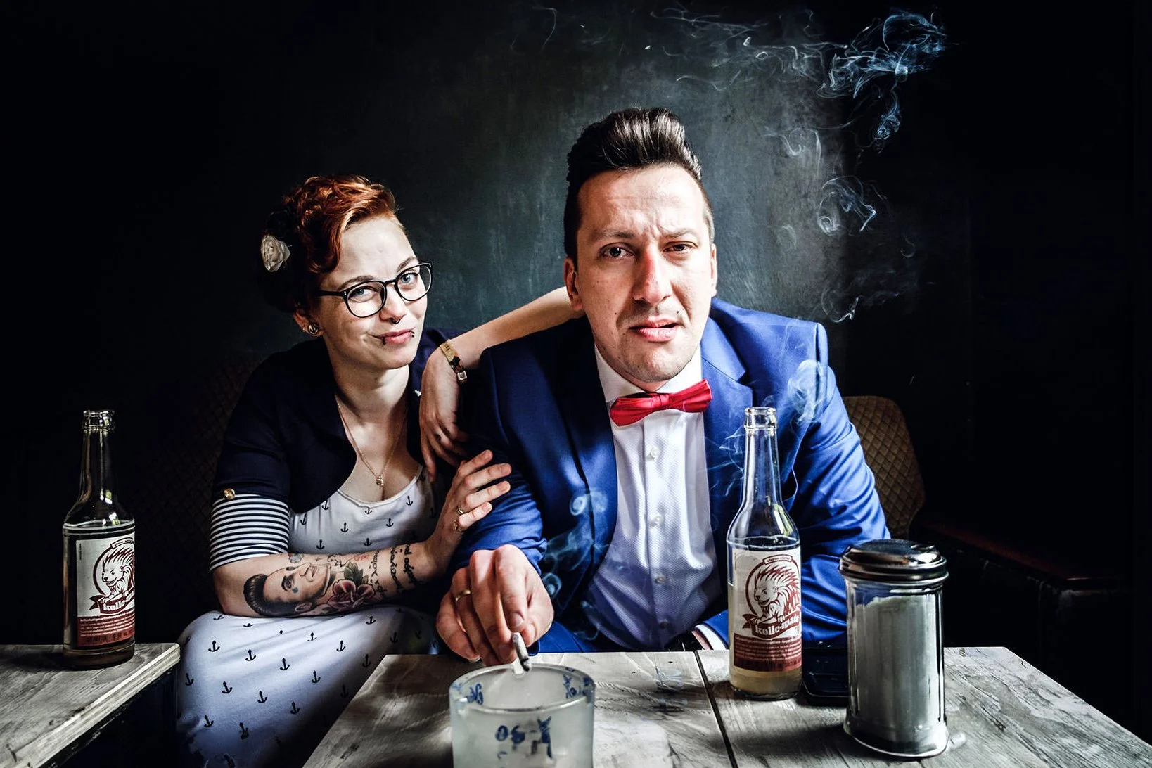 Zwei Menschen sitzen an einem Tisch in einem dunklen Raum, beide trinken Bier. Der Mann trägt einen blauen Anzug mit roter Fliege und wirkt skeptisch, die Frau hat rote Haare, trägt Brille und neckt ihn spielerisch.