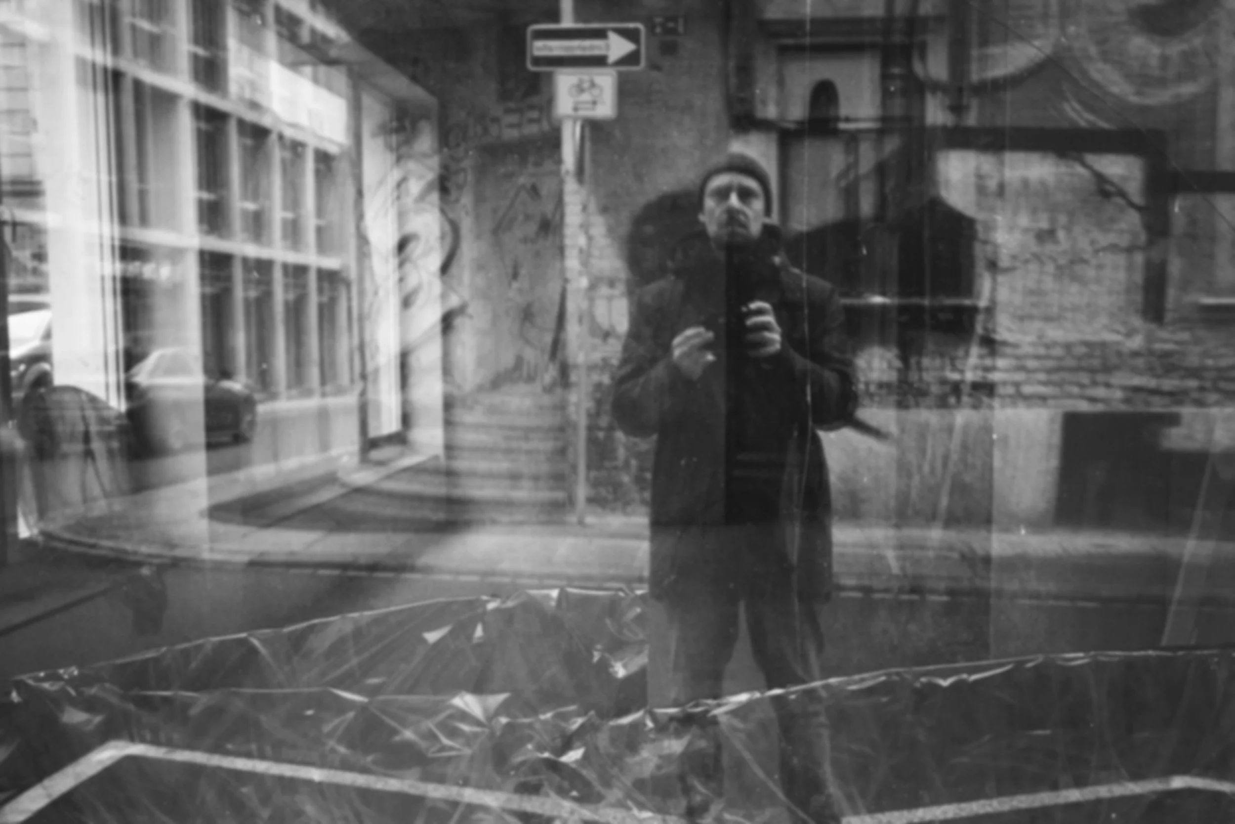Schwarze und weiße Straßenaufnahme mit reflektiertem U-Bahn-Fahrgast, der im Schaufenster einer alten Gebäudefassade fotografiert wird, umgeben von Stadt- und Fahrzeug-Reflexionen.