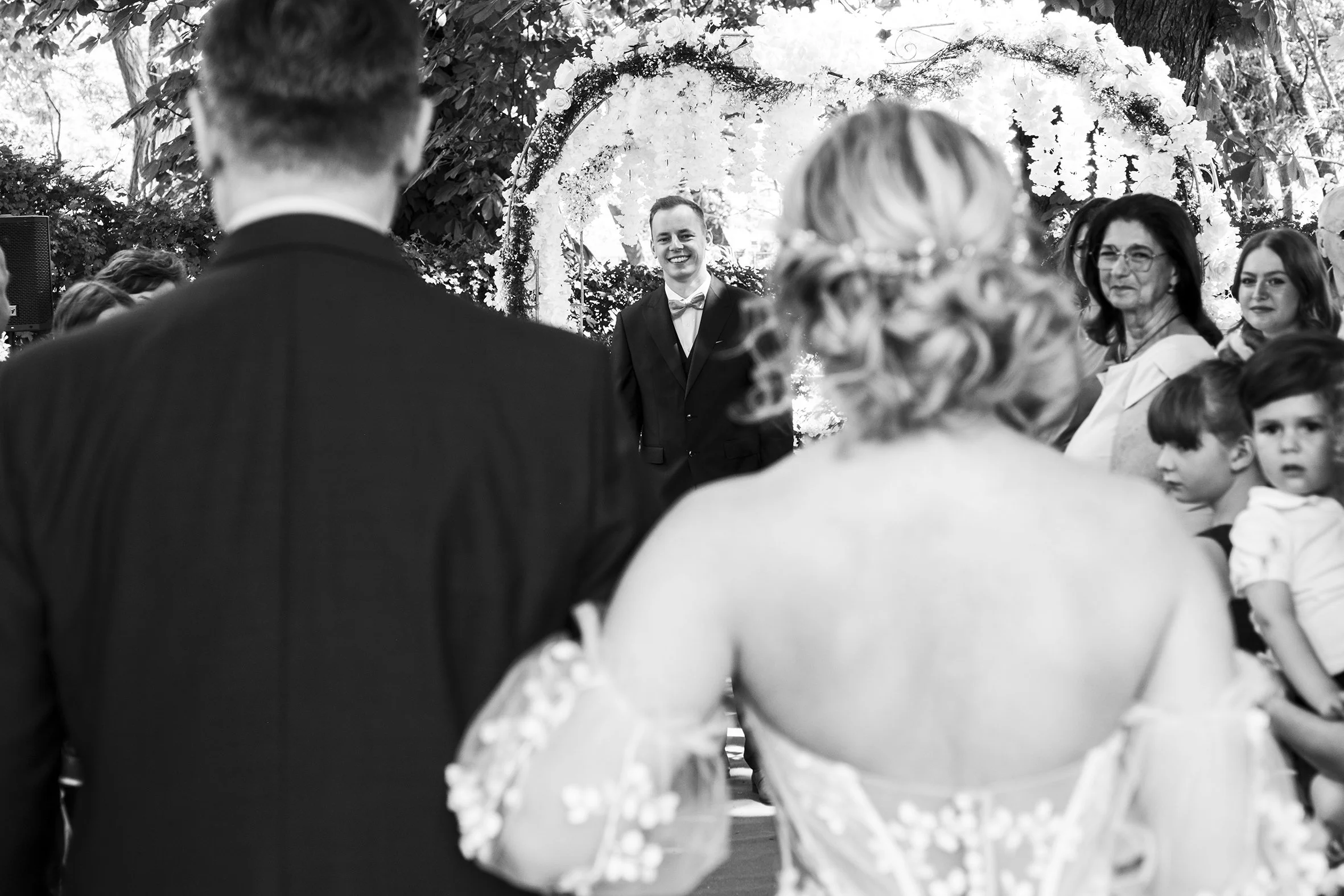 Brautpaar bei einer Hochzeit, umgeben von Gästen, im Freien unter einem Blumenbogen.