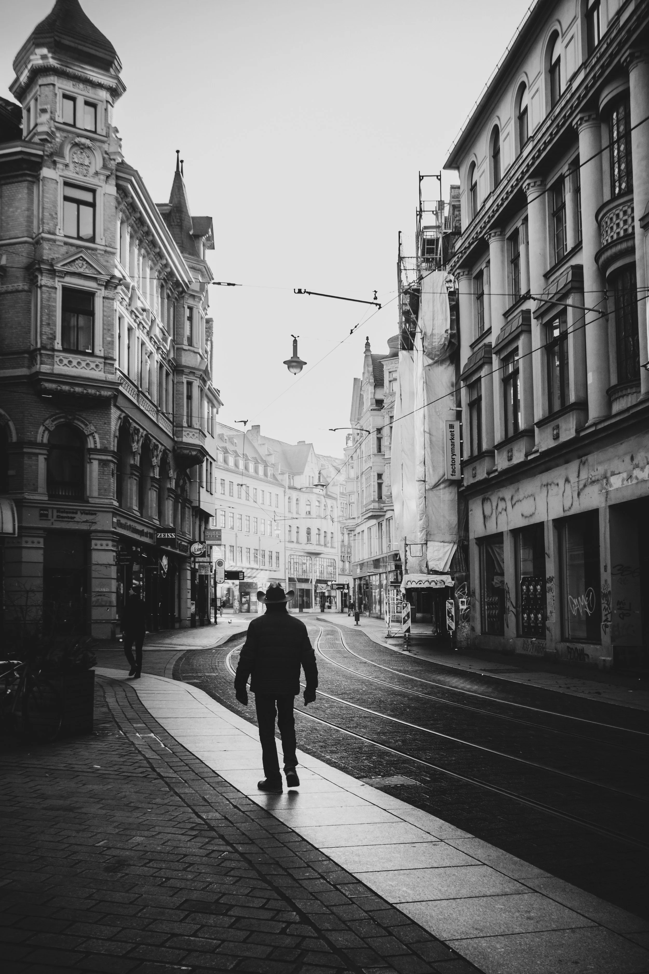 Ein schwarz-weiß Foto einer Straße in der Stadt mit mehreren Gebäuden, Straßenbahngleisen und wenigen Fußgängern, einer Person mit Hut im Vordergrund.
