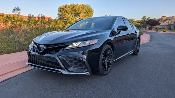 A black shiny 2020 Toyota Camry sits on a road with  blue sky and greenery behind