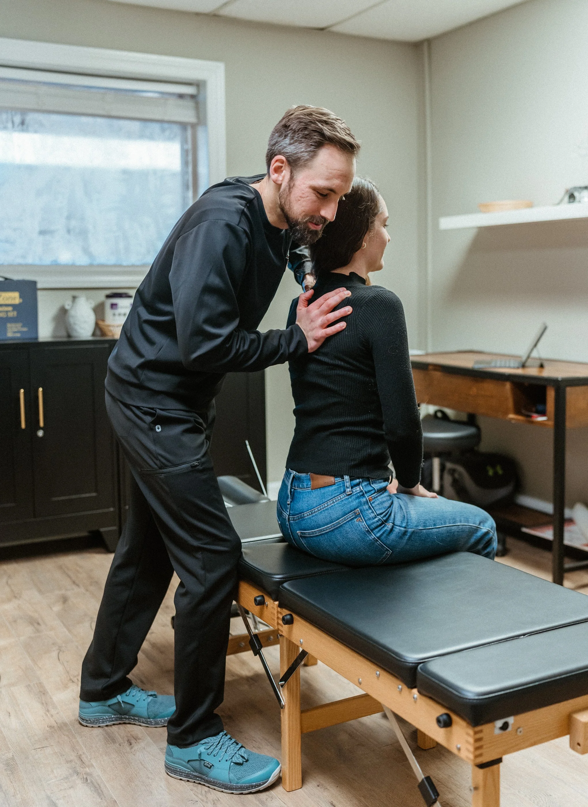 A chiropractor or physical therapist examines and applies chiropractic care to a woman sitting on a treatment table in a clinic or therapy room.