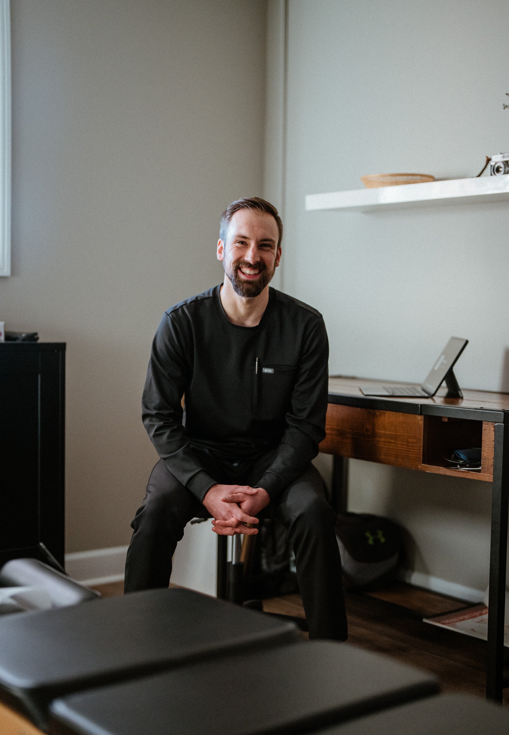 A man with a beard smiling and wearing a black jacket, seated in a room with a laptop and a camera on a shelf.