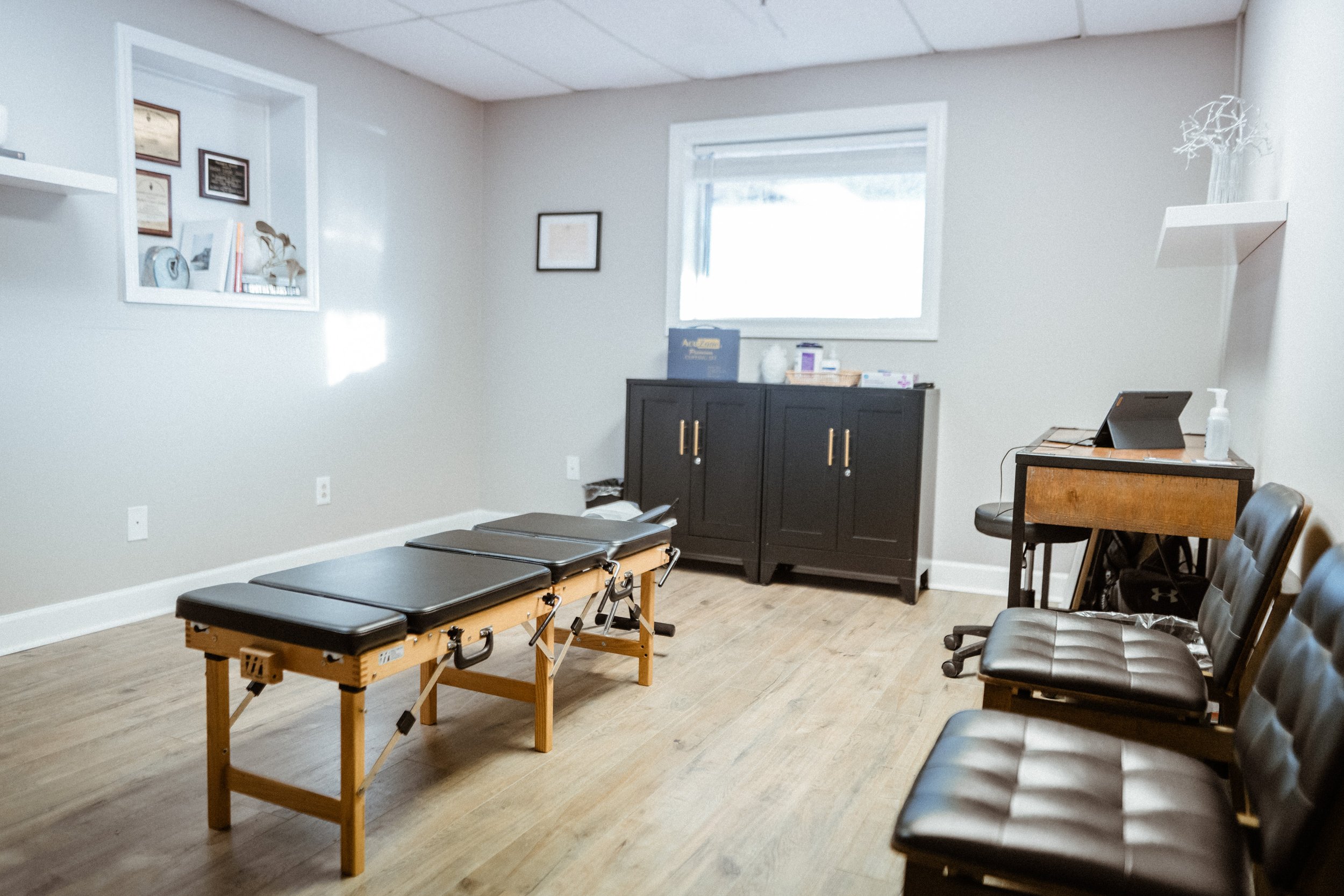 A room with light gray walls and hardwood floors, set up as a waiting or exam room. There are black leather chairs along one wall, a small table with a hand sanitizer and a monitor or tablet, a black cabinet with supplies, and a wooden examination table with a black cushioned top. The room has a window and framed artwork on the walls.