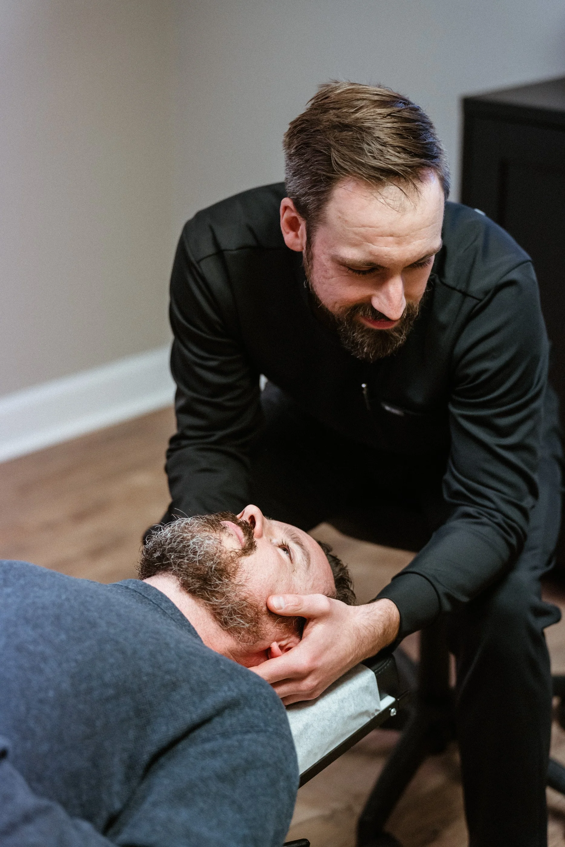 A man with brown hair and a beard is attending to a person lying on a table, possibly performing CPR or a medical procedure.