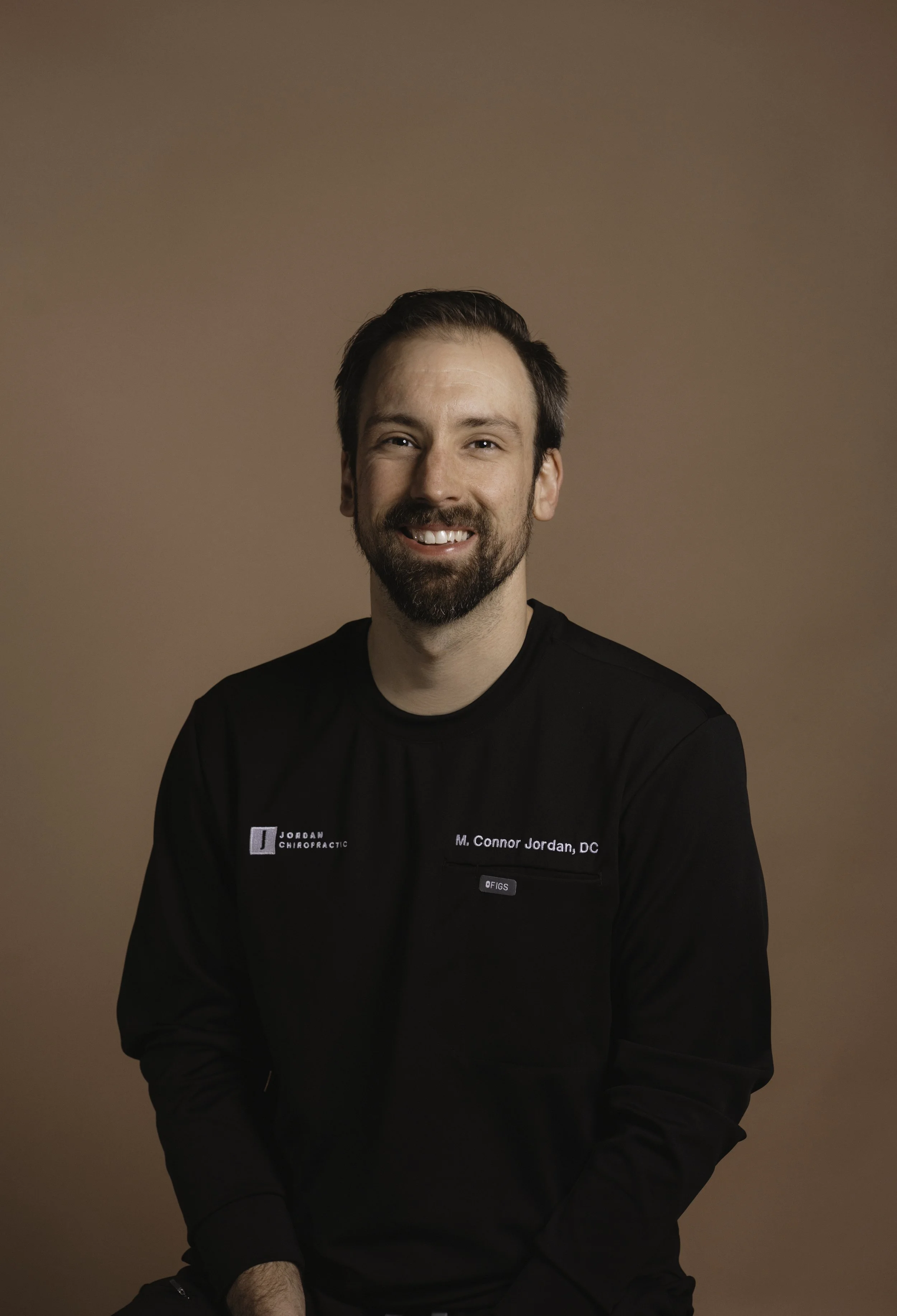 A man with dark hair and a beard, smiling, wearing a black uniform with a name tag that reads 'Mr. Connor Jordan, DC' and 'JORDAN CHIROPRACTIC,' sitting against a brown background.