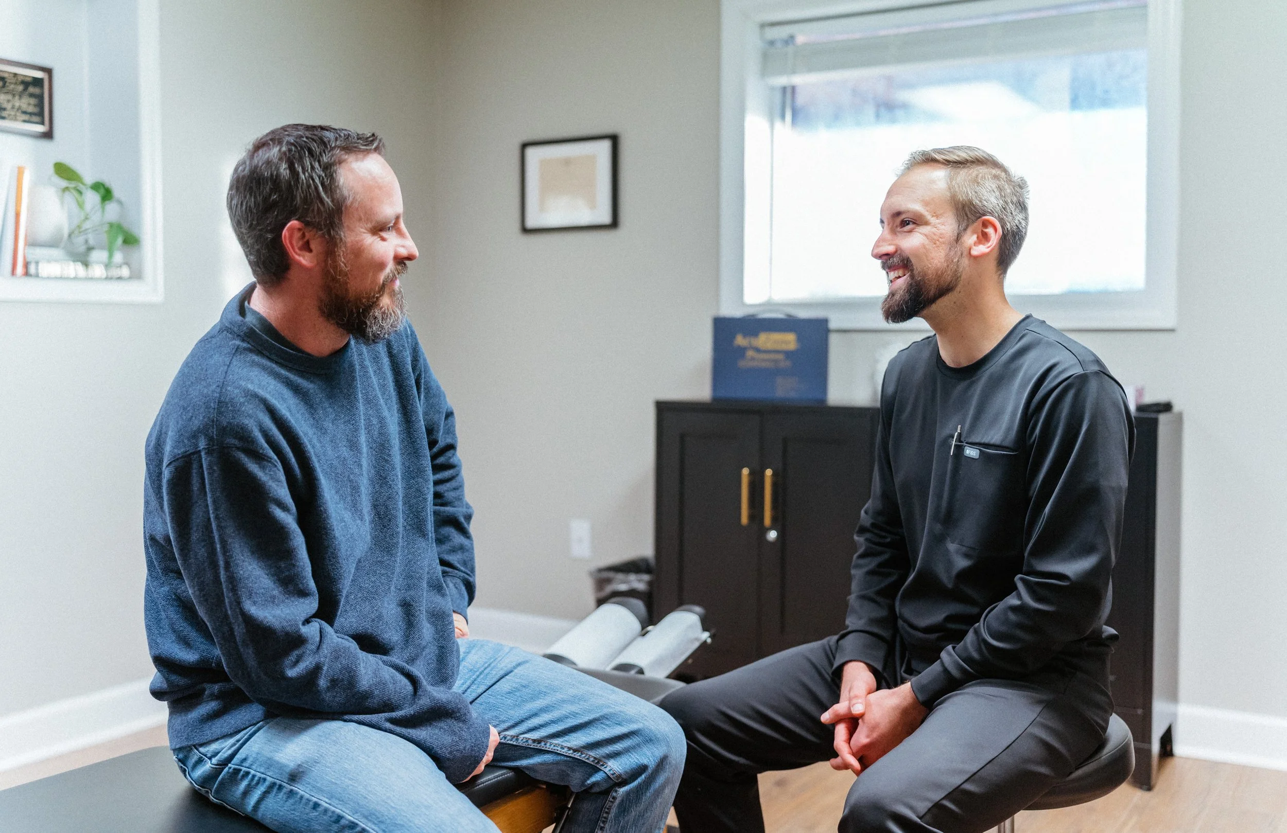 A doctor and a patient are having a conversation in a medical office. The patient is sitting on an examination table, and the doctor is sitting on a stool, facing each other and smiling.