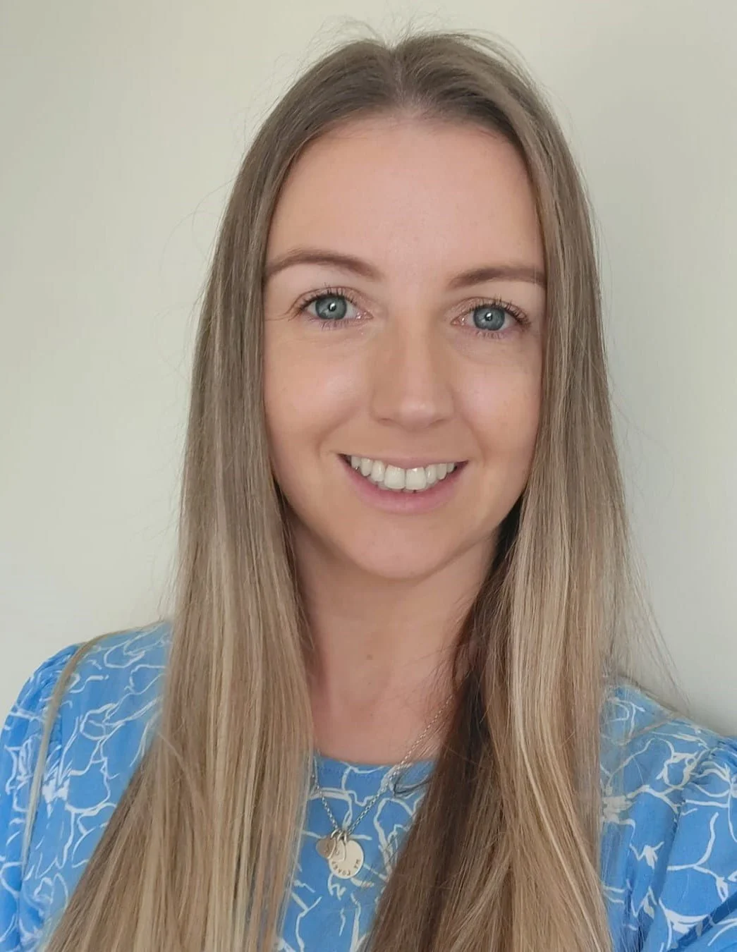 Close-up of a woman with long blonde hair, blue eyes, and a bright smile, wearing a blue patterned top and a gold necklace, standing against a plain light-colored background.