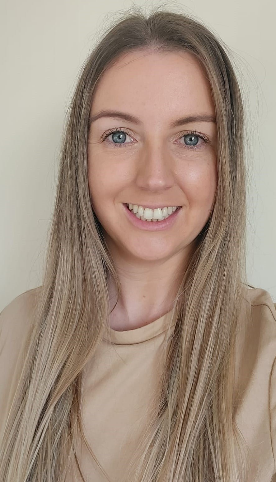 A woman with long, straight blonde hair and blue eyes smiling at the camera, wearing a beige top.