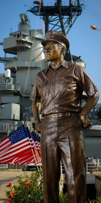 Statue of a man in military uniform with his hands on his hips, in front of a navy ship and American flags.