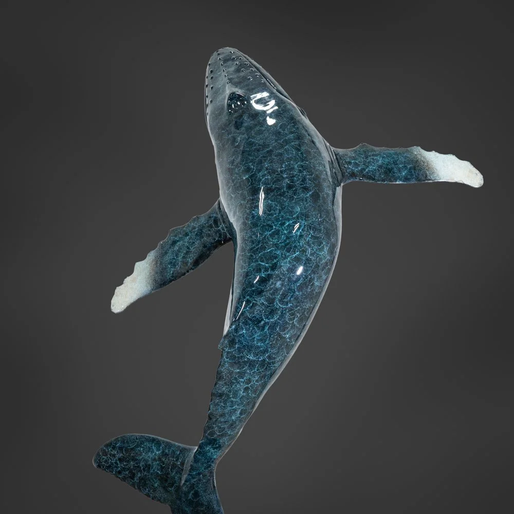 A large, bronze, patinated whale suspended in mid-air against a dark background, showing the whale's body from head to tail.