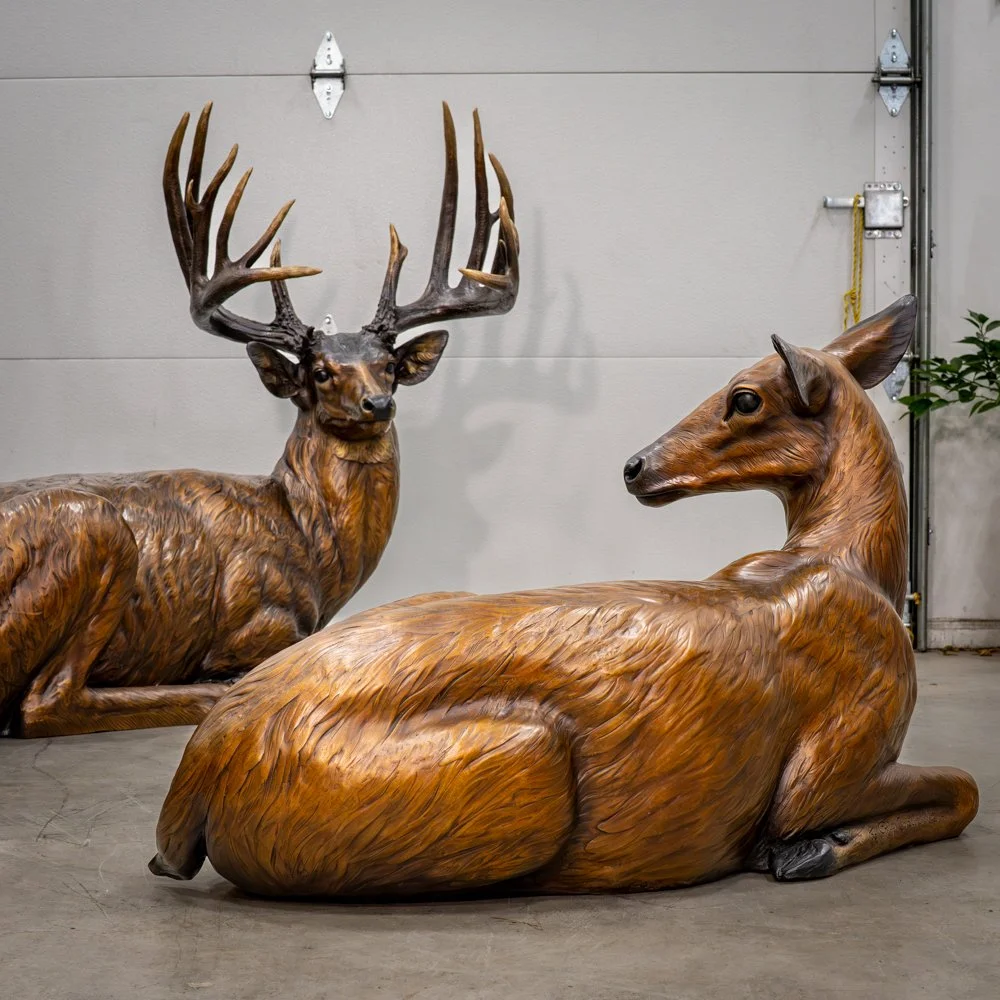 Bronze sculptures of a sitting deer with antlers and a sitting dee without antlers, with a sleek body in a garage setting.