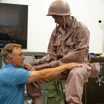 A man in a blue shirt working on a realistic sculpture of a soldier in a helmet, with the man and sculpture engaging in a collaborative pose.