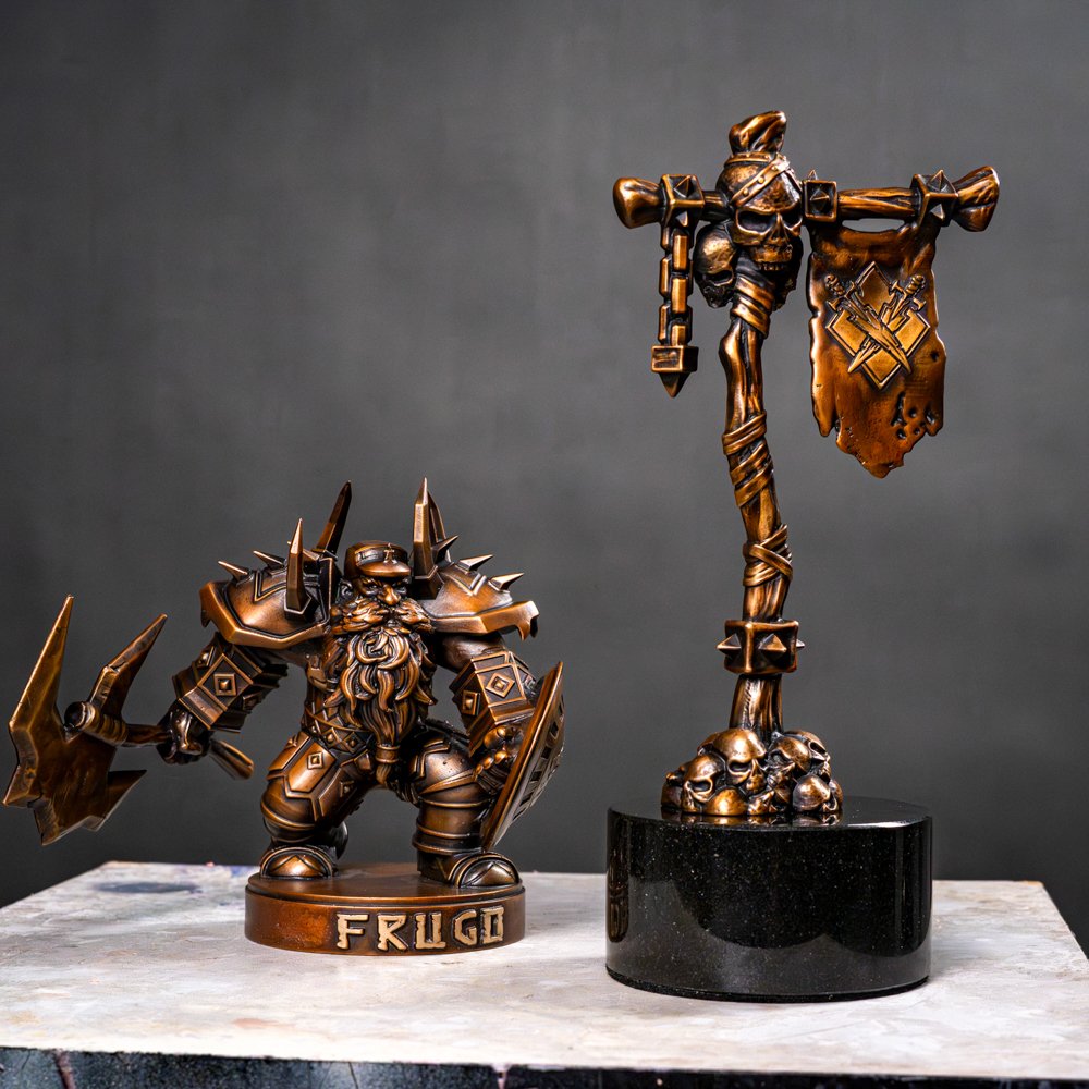 Bronze-colored figurine of a dwarf warrior with a axe and shield, labeled 'FRUGO', and a tall totem-like sculpture with skulls and flags, on a white marble surface against a dark background.