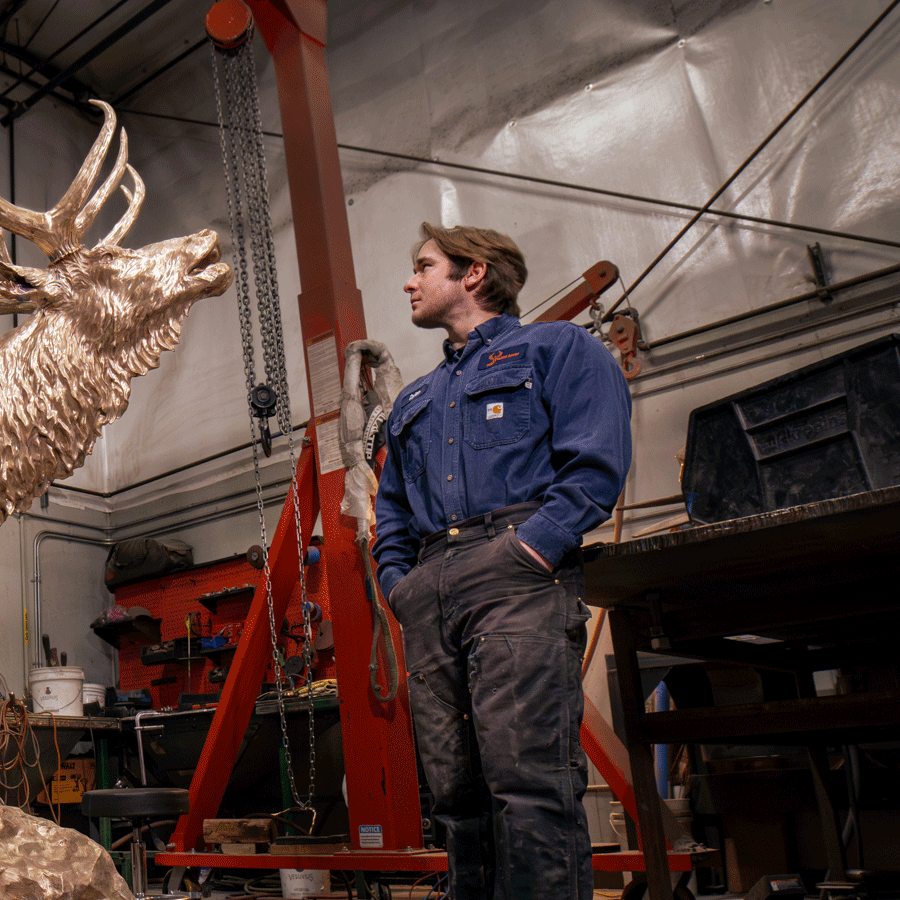 Man in blue work shirt and dark pants standing next to a large bronze elk sculpture inside a workshop.