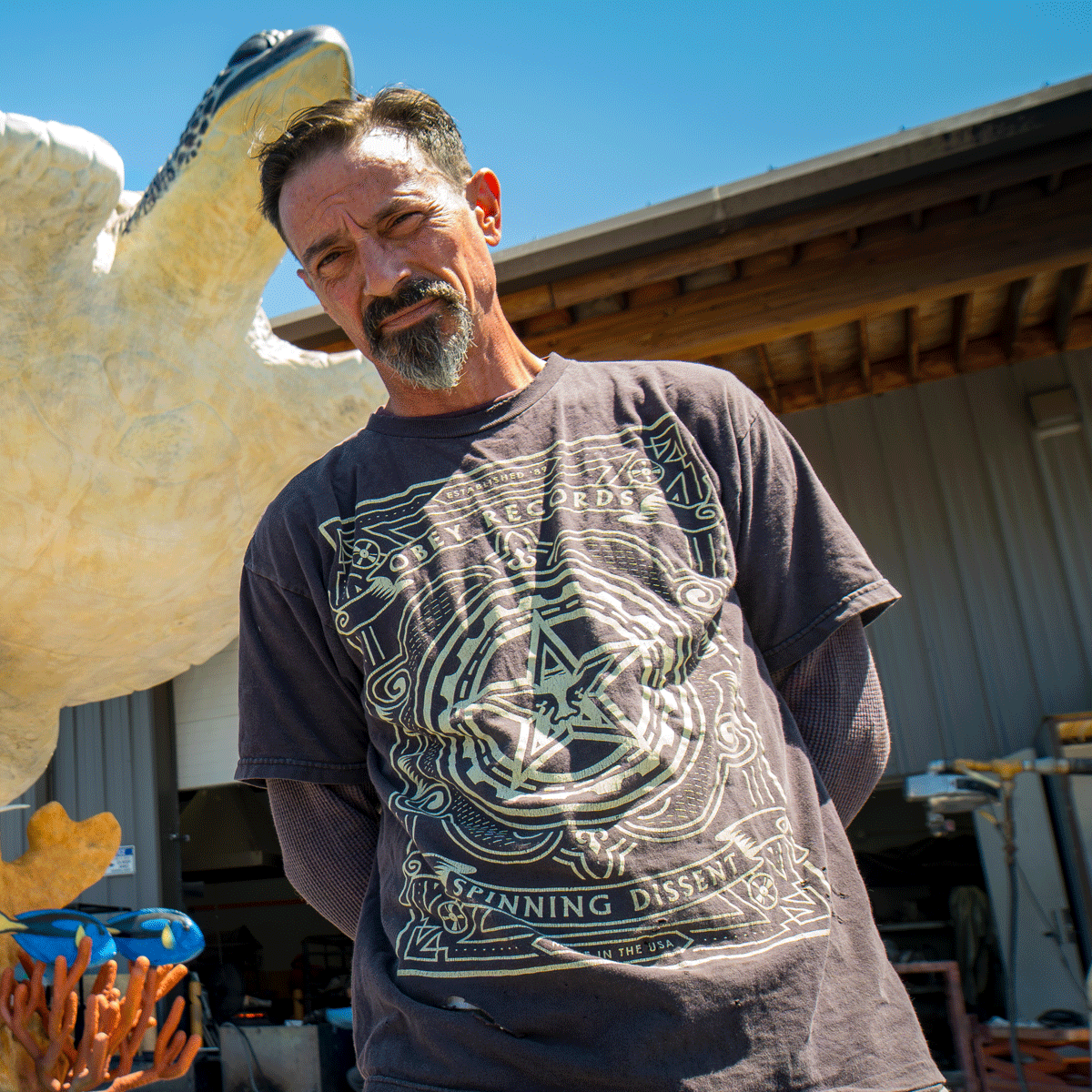 Man with a goatee and mustache standing outdoors next to a large turtle sculpture, wearing a black T-shirt with intricate graphic design.
