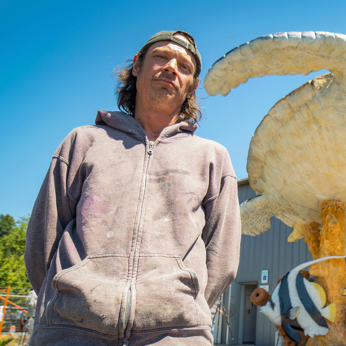 A man wearing a dirty, dusty hoodie and a backward cap stands outdoors under a clear blue sky, with a large turtle sculpture and a clownfish sculpture nearby.