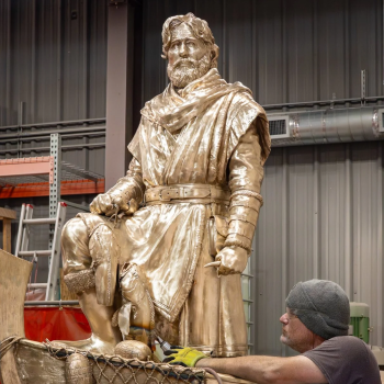 A worker is sculpting a large historical statue of a man with a beard, dressed in medieval or Renaissance clothing, in an indoor workshop.